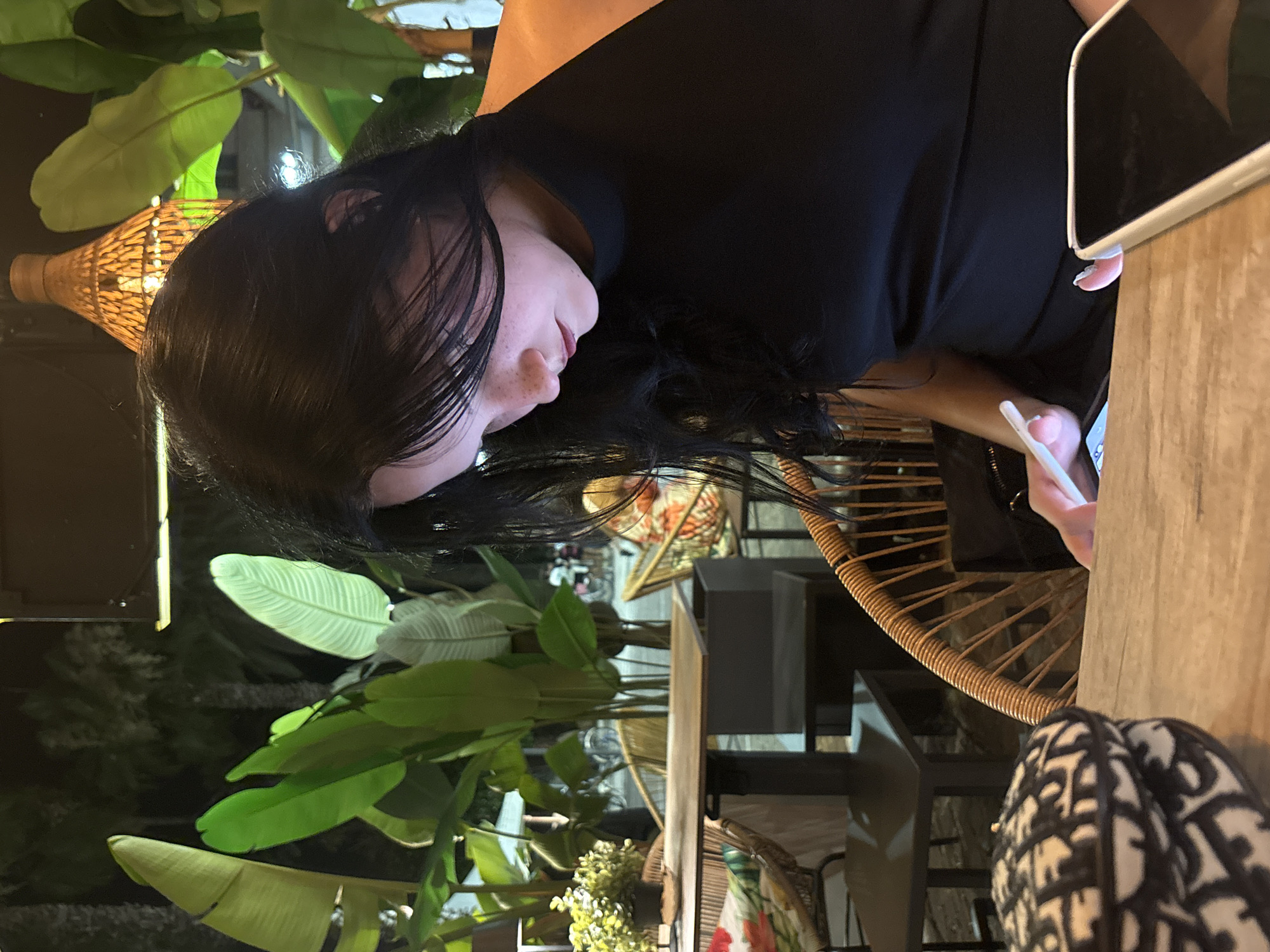 A young woman with long black hair and fair skin, wearing a black sleeveless top, is seated at a table in an outdoor restaurant setting. She is looking down at her phone, her face illuminated by the screen's light. Her hair partially obscures her face, and she has freckles. To her left, on the wooden table, is a black and white patterned round object, possibly a hat or bag. In the foreground, another phone with a white case is visible. The background features lush green tropical plants, wicker chairs, and other patrons at tables, suggesting a vibrant, possibly nighttime, atmosphere in San Benedetto del Tronto, Italy. A woven lamp hangs from above, casting a warm glow.