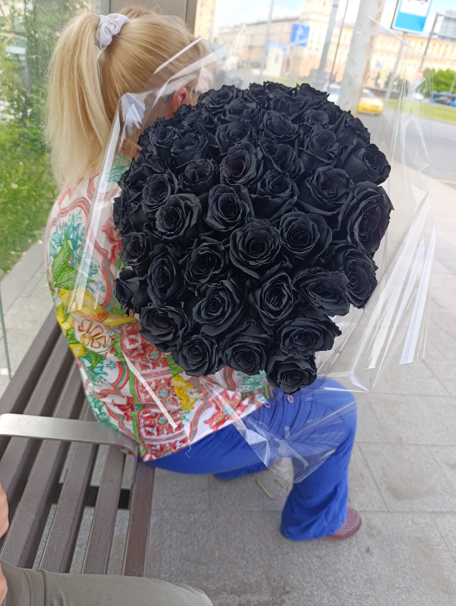 A person with blonde hair tied in a ponytail sits on a slatted brown bench outdoors, holding a large bouquet of black roses. The person is viewed from the back and side, wearing a vibrant, colorful patterned shirt adorned with fruit motifs, notably lemons, and bright blue pants, with pink slip-on shoes visible. The dense bouquet of black roses is wrapped in clear, transparent plastic, occupying a significant portion of the foreground.
The setting appears to be an urban environment in Moscow, Russia, likely a sidewalk or bus stop area. Through the transparent plastic wrapping and in the background, blurry city elements are visible, including buildings, what seems to be a street, and a yellow vehicle. A blue sign, typically indicating a bus stop with a white bus icon, is faintly discernible in the distance, further suggesting a public transit area. The bright, even lighting indicates it is daytime. The person is seated, seemingly resting or waiting, while prominently displaying the striking, dark roses. No explicit text is clearly visible other than the bus icon on the distant sign.