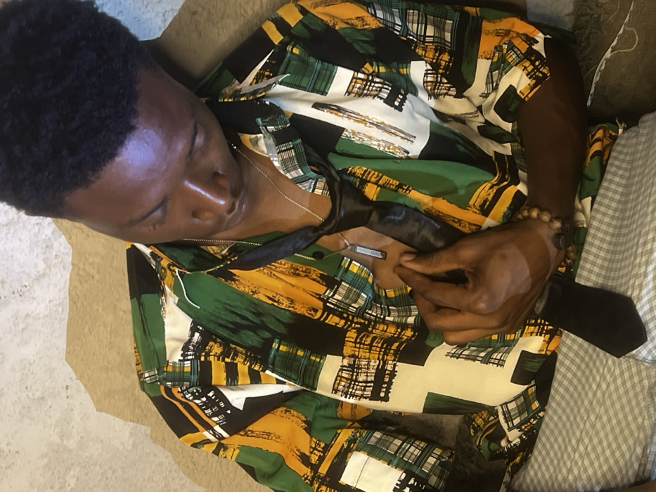 A young Black male is depicted from an elevated perspective, seated or leaning against a surface. His head is tilted downwards and slightly to his right, with eyes cast down or closed. He wears a short-sleeved collared shirt featuring an abstract pattern of green, yellow, black, and white brushstrokes. An untied black necktie is draped around his neck, resting against his chest. A silver-colored chain with a rectangular pendant hangs around his neck. His right hand is positioned on his chest, lightly gripping the black tie. A bracelet made of wooden beads is visible on his right wrist. He is wearing light-colored, possibly checkered, lower garments. The immediate background behind his head and left shoulder is a light tan, textured surface, while a darker, indistinct textured surface is visible to his right. The scene occurs in Bar Gada, Nigeria.