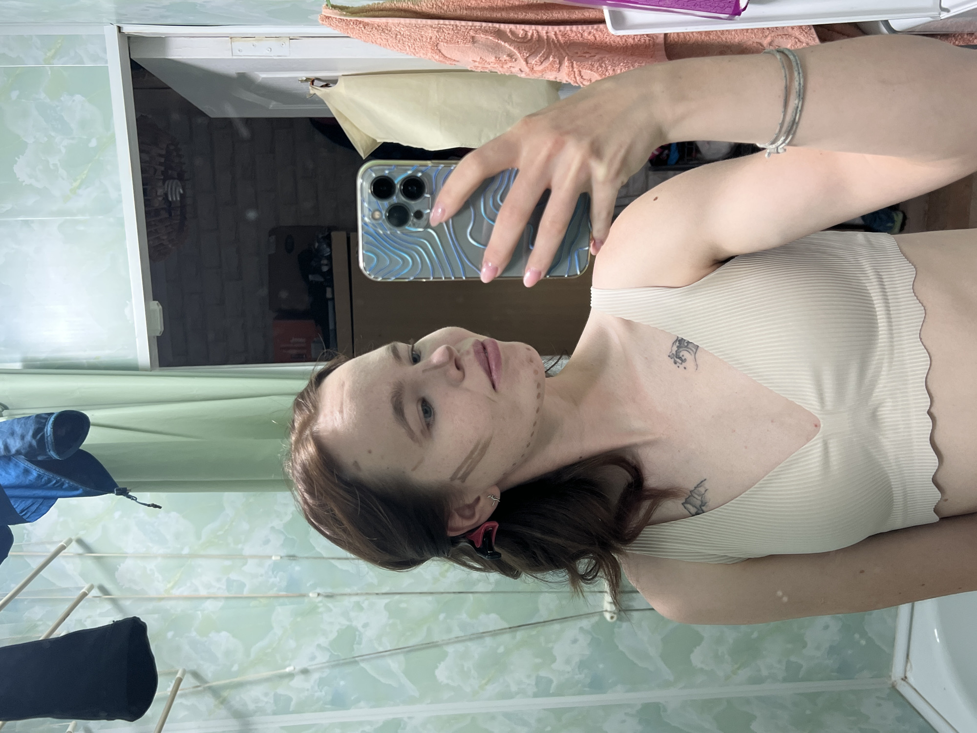 A woman is taking a selfie in a bathroom. She is holding an iPhone with a textured blue case. She is wearing a light beige sports bra. There are lines drawn on her face, potentially for contouring makeup. She has brown hair with a pink clip visible. The bathroom has green tiled walls and a clothes drying rack with blue clothing hanging on it. Behind her, a mirror reflects a portion of another room. The location is Chaikovsky, Russia.