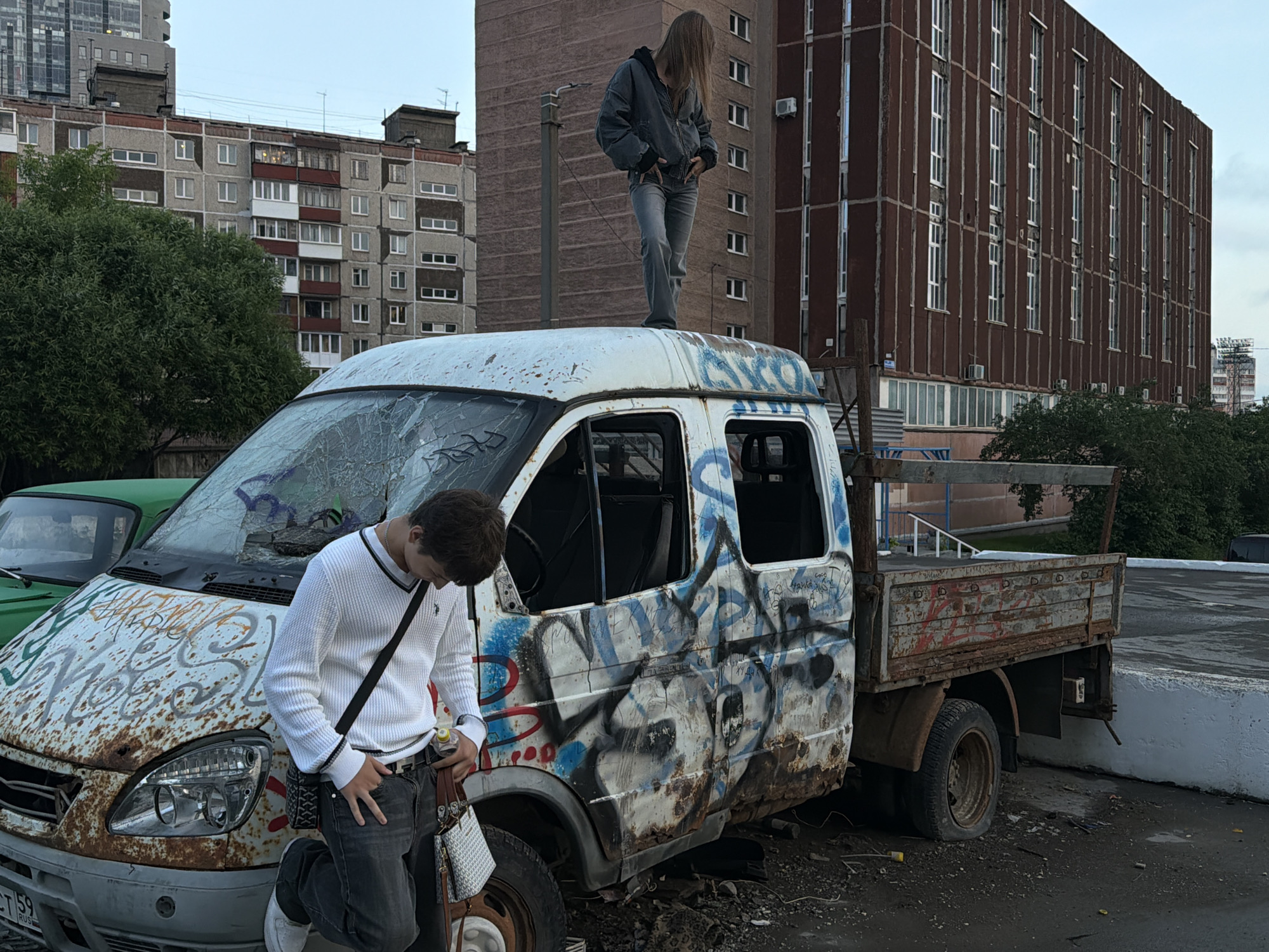 A young person with long, blonde hair stands on the roof of a rusty, graffiti-covered white van in what appears to be an urban environment. They are wearing a dark jacket and grey jeans. In the foreground, another young person, wearing a white sweater with black trim, dark jeans, and sneakers, leans against the van. They are holding a bottle of water and have several bags. The van is parked on a paved surface, possibly a street or parking lot, with debris scattered around it. In the background, several apartment buildings and a larger, modern building are visible. The sky is overcast. The presence of graffiti on the van and the general state of disrepair suggest an industrial or neglected area. Based on the context of other images, this scene is located in the city of Perm, Russia, likely in an industrial or residential area with evidence of urban decay.