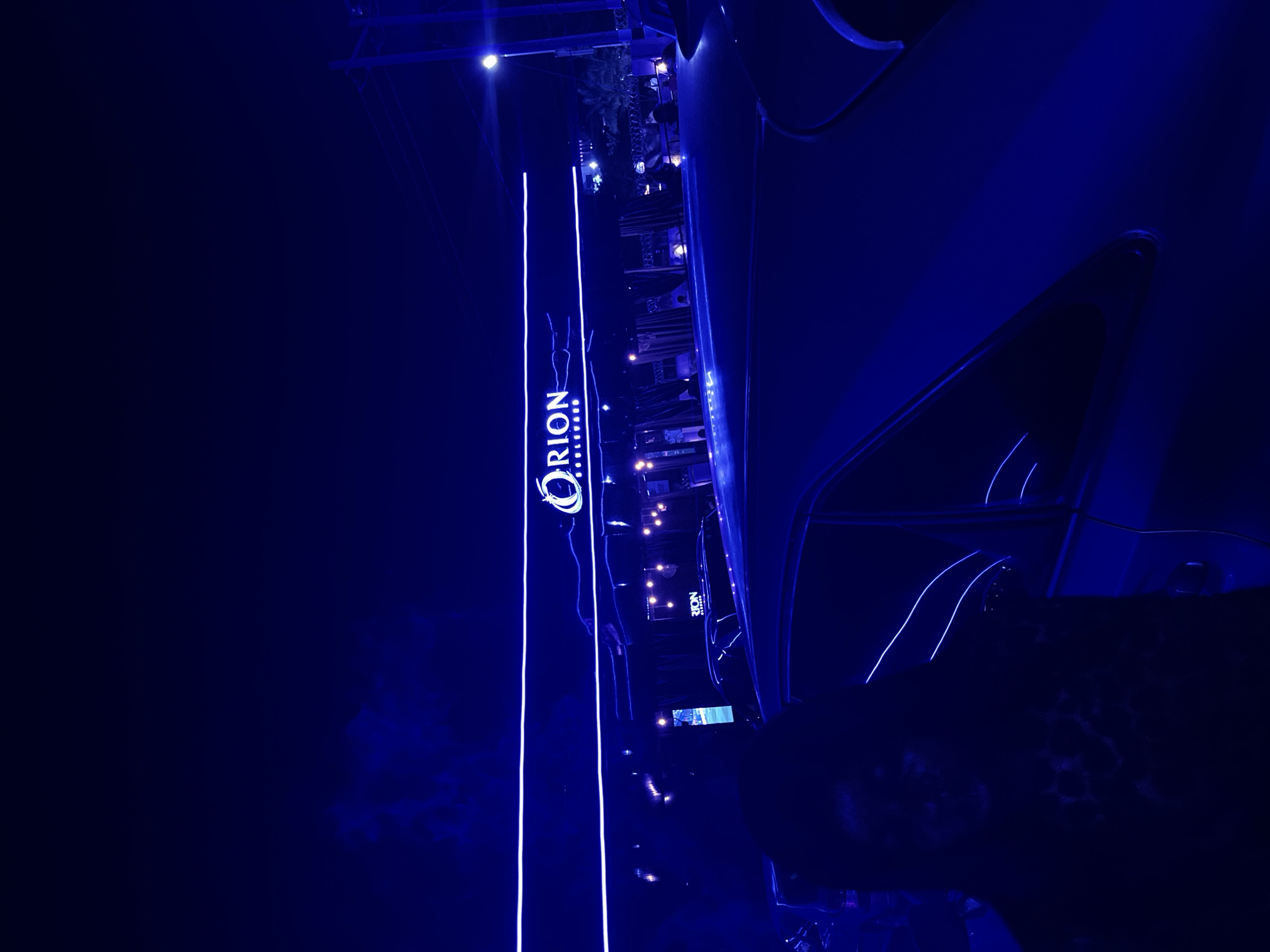 A person is standing next to a car at night in Abuja, Nigeria. A building with a neon sign that reads "ORION" is visible in the background. The scene is illuminated by blue neon lights, giving it a dark and moody atmosphere.