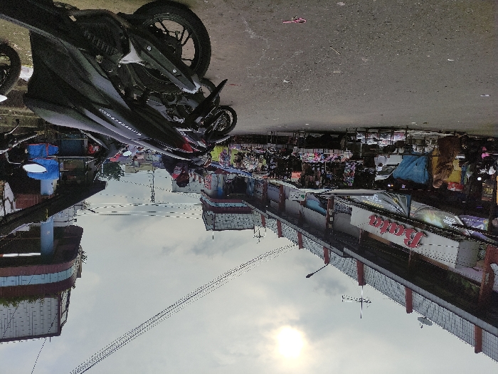 A black motorcycle is parked on the street in front of a building, partially obscured by the motorcycle's large, silver, front tire. The front end of the motorcycle is pointed towards the right side of the image. The street is made of asphalt. The camera is positioned on the ground, looking upwards, so the buildings in the background appear upside-down. The buildings have red and white trim. There are many people walking in the distance, obscured by the motorcycle and the perspective of the image. The sky is cloudy and the sun is shining in the top right corner. The building in the background has a red sign that says "Raja" on it. The scene is located in Kecamatan Lawang, Indonesia, at latitude -7.83, longitude 112.697.