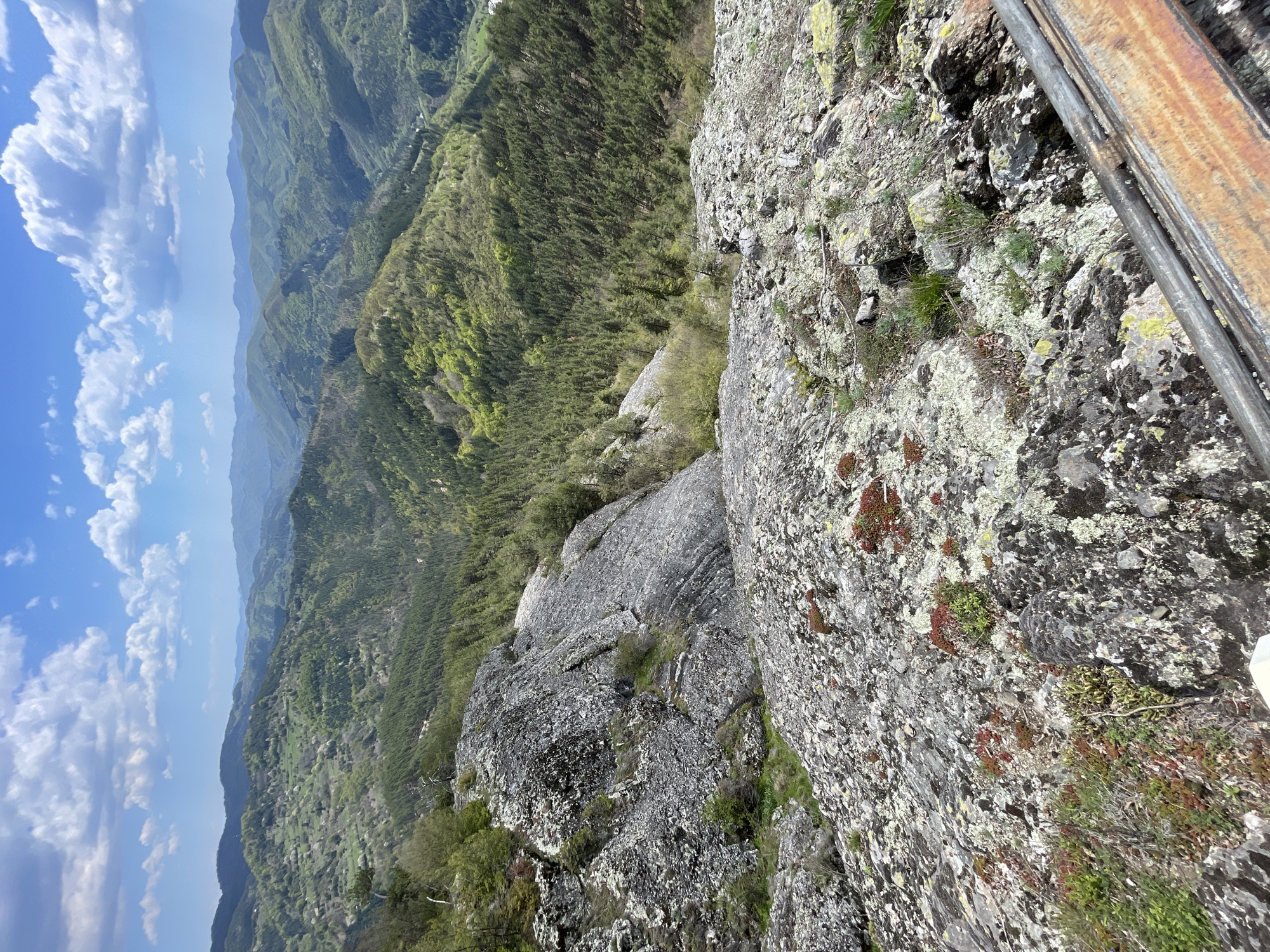 The image shows a rocky cliff overlooking a mountainous, forested landscape. In the background, a blue sky with scattered white clouds is visible. The foreground features gray rocks covered in lichen and small patches of vegetation. A metal railing is visible on the right side of the frame. The view is from the "The Eye" observation deck in Gradat, Bulgaria.