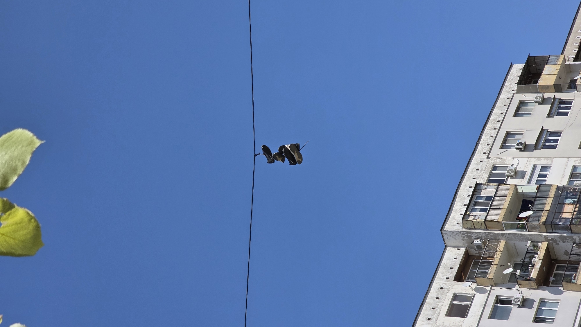 A low-angle shot shows a pair of shoes hanging from a black cable stretched across a clear blue sky. The cable extends vertically from the bottom center of the frame to the top center. The shoes are dark-colored and appear to be sneakers or athletic shoes. To the right of the cable, a white multi-story building with windows, balconies, and air conditioning units is visible. To the left of the cable, the edge of a leafy green tree branch is present. The building suggests an urban environment, potentially in Blagoevgrad, Bulgaria, though this is difficult to confirm without additional contextual cues. There are no people visible in the scene.