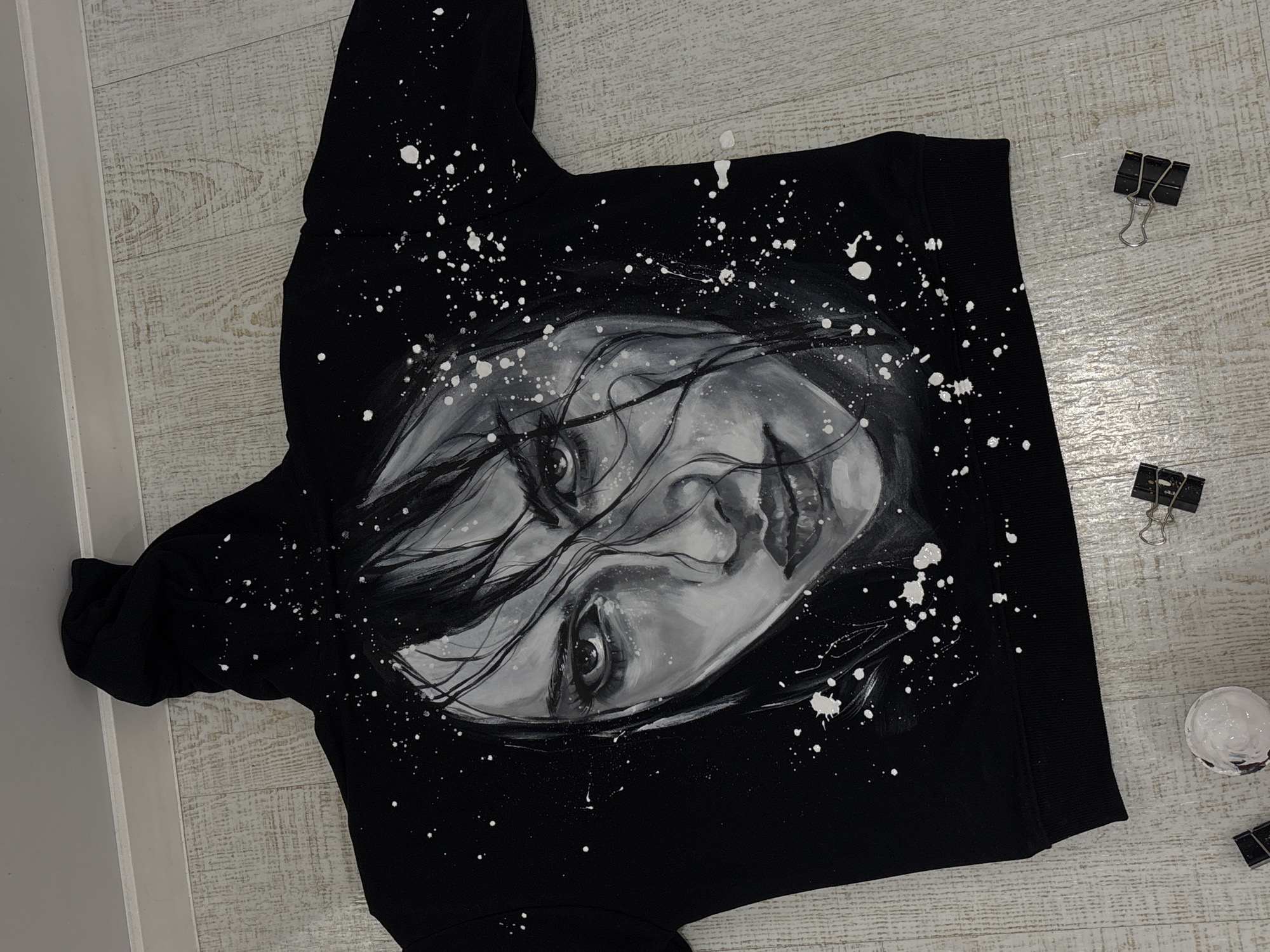 A black hoodie with a monochrome print of a woman's face is laid out on a light-colored wooden floor. White paint splatters are scattered around the print and on the hoodie. Two binder clips and a small jar with a white substance are arranged around the hoodie on the floor. The hoodie is not in Krasnoyarsk, Russia.