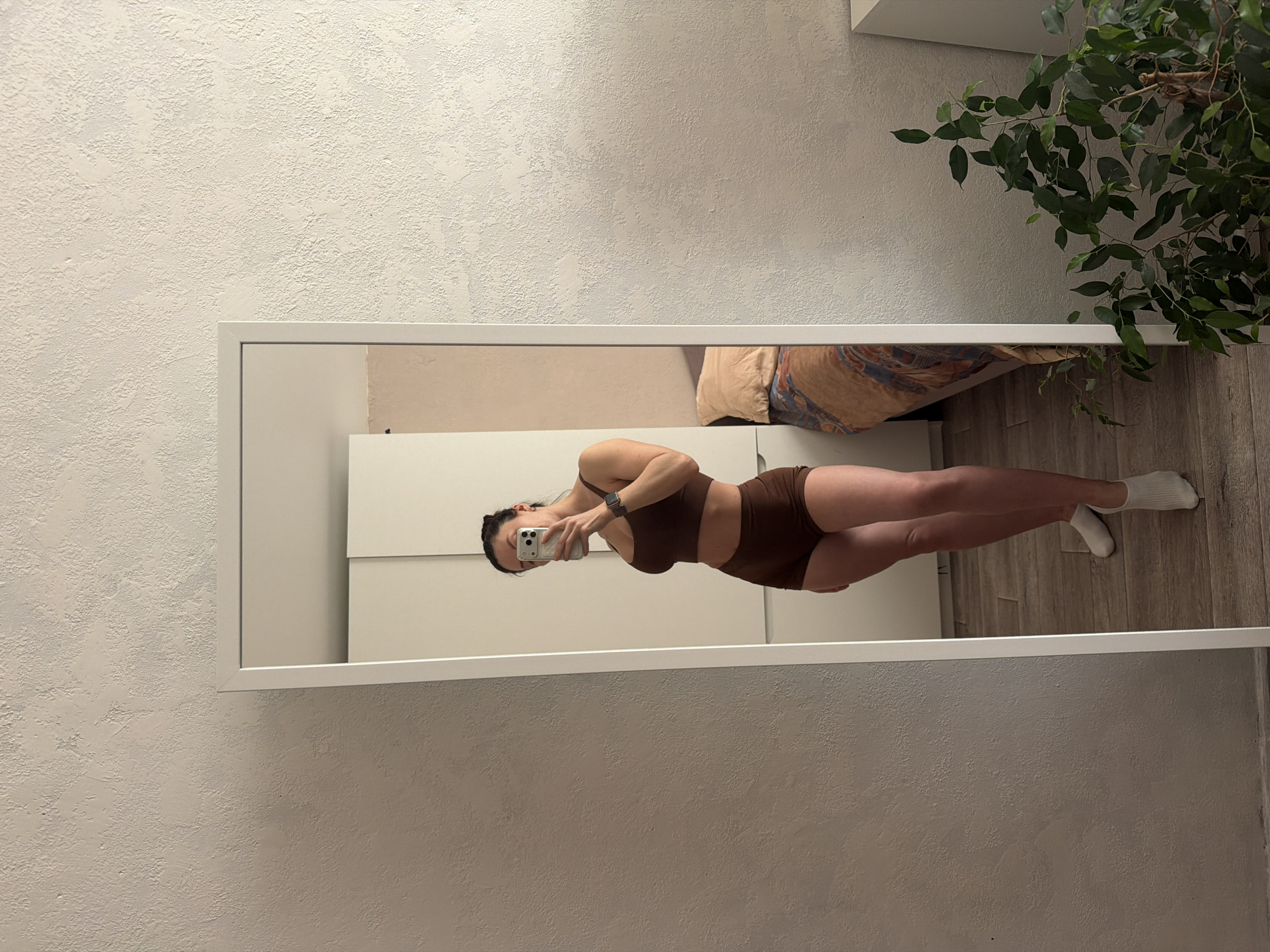 A person stands in front of a full-length mirror, holding a smartphone up to take a picture of themself. The person is wearing a brown two-piece outfit and white socks. The mirror is positioned against a textured off-white wall. Reflected in the mirror is a bedroom setting with a beige bed and a white wardrobe. A potted plant is located to the right of the mirror. This photo was taken inside of a home, potentially in the city of Perm, Russia.