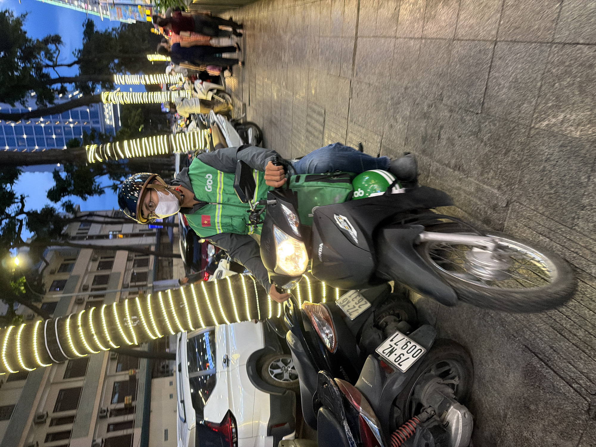 A man wearing a white face mask, glasses, and a dark helmet with a red star emblem is riding a black motorcycle. He is dressed in a green vest with a "Grab" logo and blue jeans. The motorcycle has a green delivery bag on the front. He is on a sidewalk next to a street lined with trees decorated with strings of yellow lights. In the background, there are buildings, other motorcycles, and people walking. The license plate on the motorcycle behind him reads "79-N2 009.71". The scene appears to be in Nha Trang Ward, Vietnam, given the Grab delivery service and Vietnamese license plate.