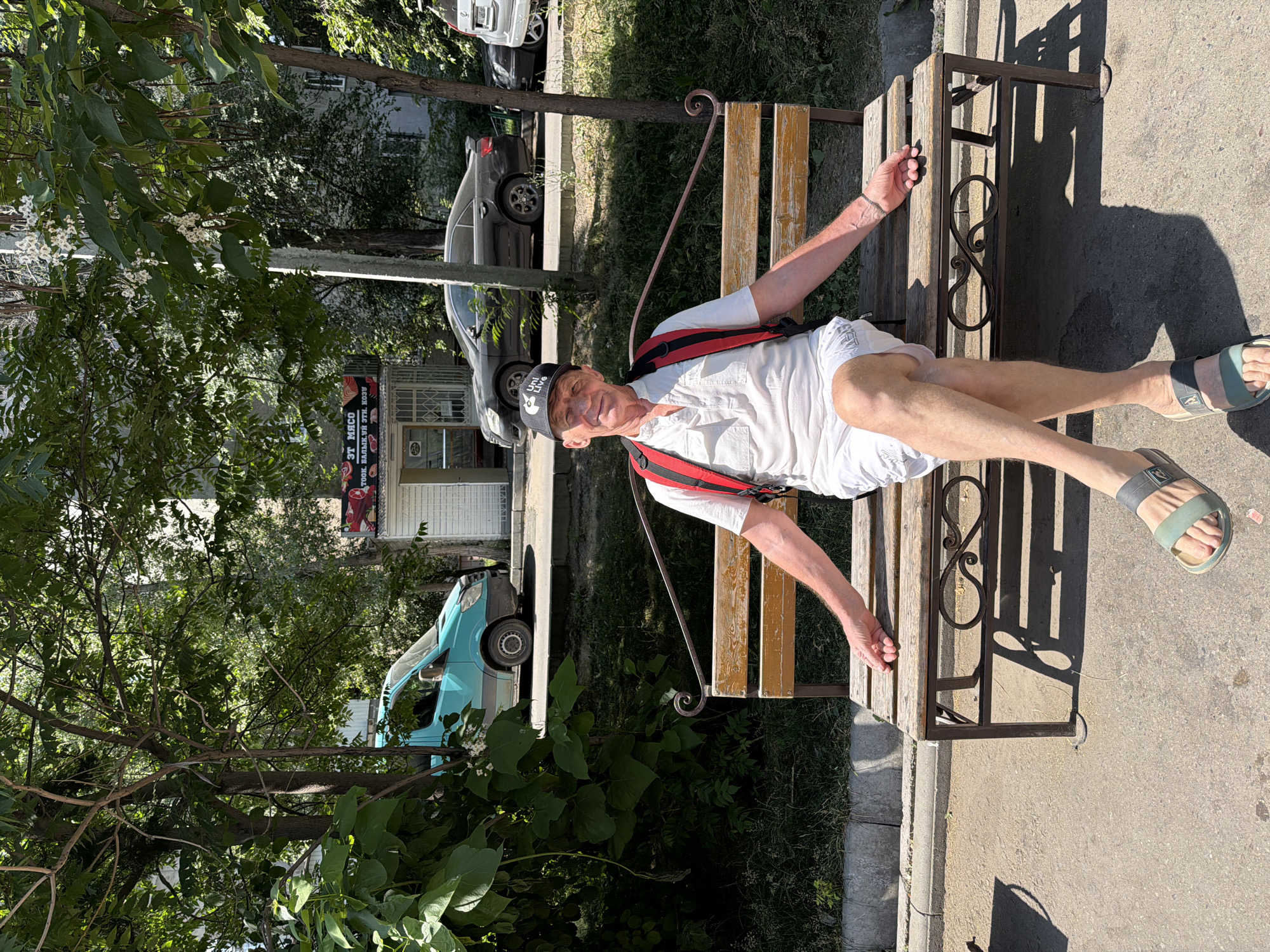 A man is sitting on a park bench outdoors in the daytime. He is a Caucasian man with light skin and short gray hair, wearing a white short-sleeved collared shirt, white shorts, a black baseball cap with a white logo, and green and gray sandals. He has a red backpack on. He is seated with his legs crossed and his arms resting on the bench. The bench is made of wood and metal and is positioned on a paved surface next to a grassy area. In the background, there is a turquoise van, a dark sedan, and a building with a sign that reads "ЭТ МЯСО." Trees and foliage surround the scene.