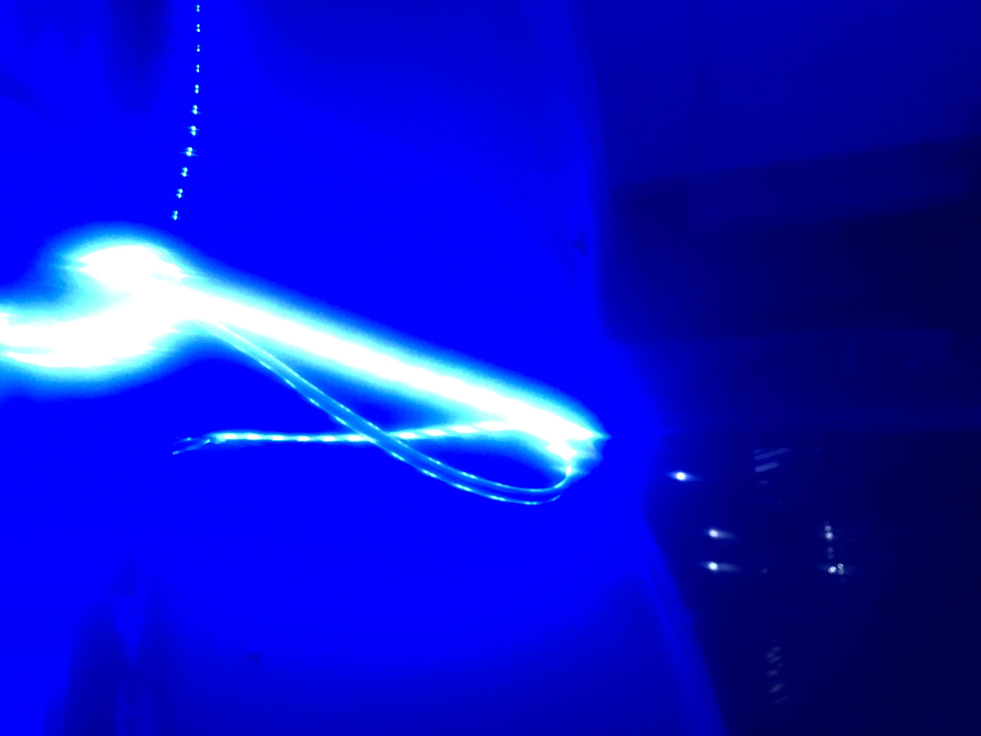 The image depicts a dark, blurry scene dominated by bright blue and white light streaks, suggesting long exposure photography or movement of light sources. The background is a deep, saturated blue, with some darker areas at the bottom suggesting a surface or floor. In the upper right portion of the image, a series of small, evenly spaced blue lights are visible, forming a line that trails off into the distance. A prominent, thick streak of bright white and blue light curves across the center of the frame from the top left to the bottom right, with thinner, more intricate light trails wrapping around it, possibly indicating movement of a light-emitting object or person. The overall impression is abstract and dynamic, with no discernible landmarks or specific environmental details to place it in Howayi, Nigeria, or any other specific location. The image is highly processed, with the lights appearing to be the sole focus of the composition, obscuring any contextual information.