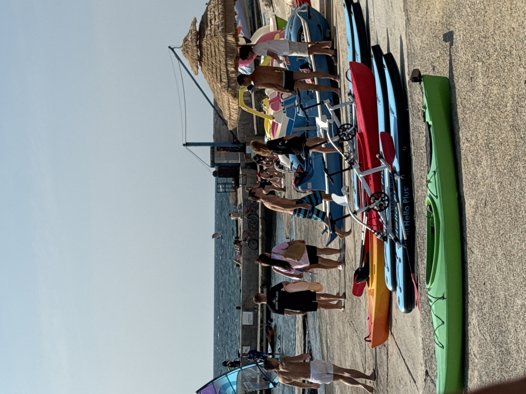 The image depicts a sunny day at a waterfront location, likely a beach or pier, with several people and various watercraft. In the foreground, a bright green kayak rests on a concrete surface. Behind it, a red kayak and a blue kayak are also visible, some with paddles and what appear to be wheeled stands. Further back, a group of people are gathered near a dock. A man in white shorts and no shirt is walking away from the camera. Another man in a black t-shirt with "STONE ISLAND" printed on it is walking with a large inflatable ring. A woman with long dark hair is carrying a beige bag. Several other people are standing or walking around the area. In the background, the sea stretches to the horizon under a clear blue sky. There are several boats, including a pedal boat with a yellow canopy, and a windsurfing sail. Some bicycles are parked on the dock. The overall atmosphere suggests a recreational area, possibly a rental spot for water sports. The location is described as Unknown, Unknown.