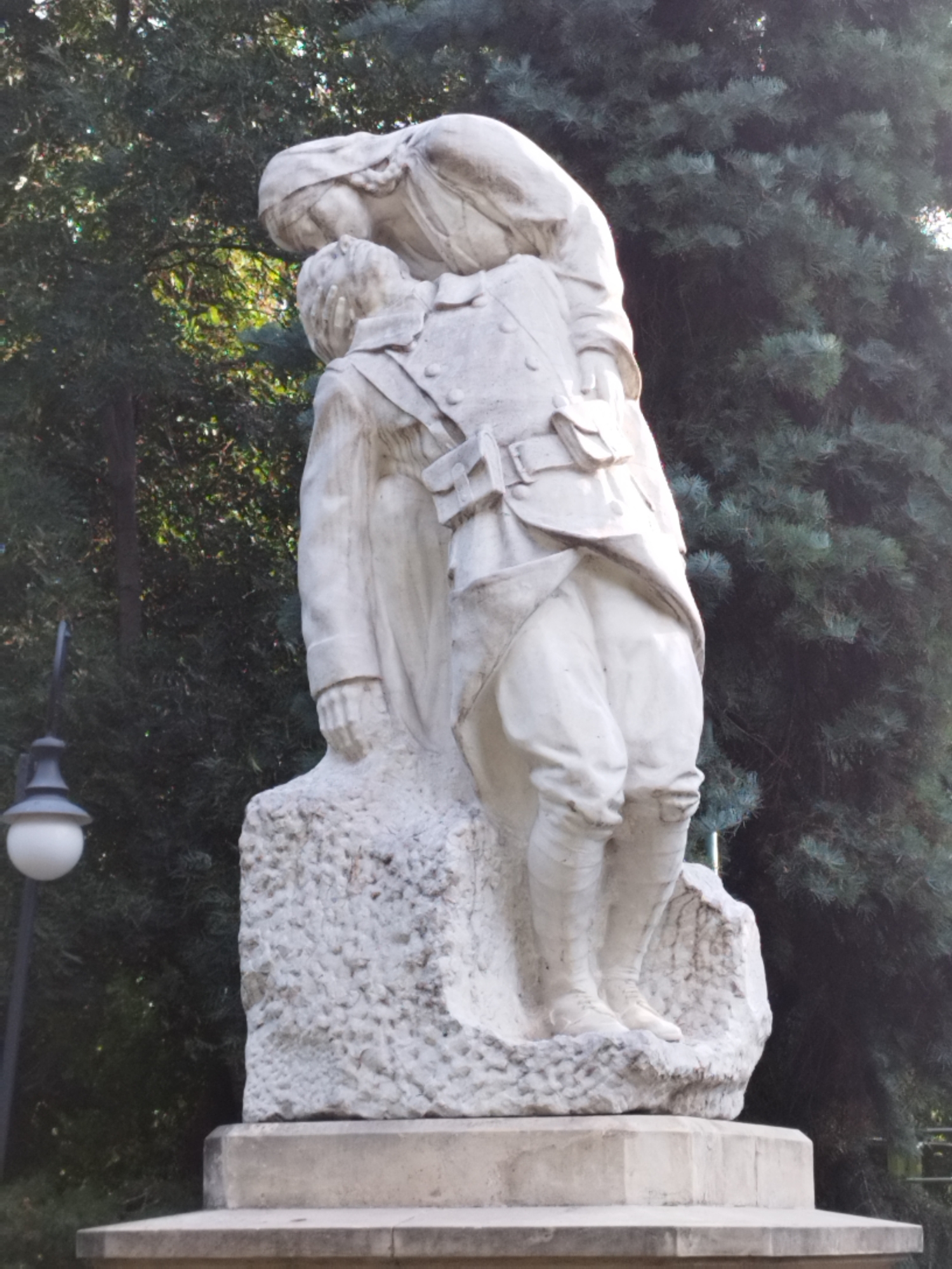 This is a vertical, medium shot of a white stone sculpture of two people in front of a dark evergreen tree. The sculpture depicts a soldier in uniform and a woman in a headscarf. The soldier, seen from the waist down and partially from the chest up, is leaning forward and appears to be receiving a kiss from the woman. The woman is bending down, embracing the soldier, and her head is tilted to kiss his cheek. The soldier is wearing a military-style uniform with a jacket, pants, and boots, and has a bandolier across his chest. The sculpture is mounted on a rough, stone base, which is then placed on a concrete pedestal. To the left of the sculpture, a black lamppost with a white globe is visible. The overall impression is one of a tender farewell or remembrance.
This sculpture is located in Bucharest, Romania. It is known as "The Kiss of the Soldier" or "The Soldier's Farewell" and is part of the monument dedicated to the heroes of the First World War. It is situated in the Carol I Park.