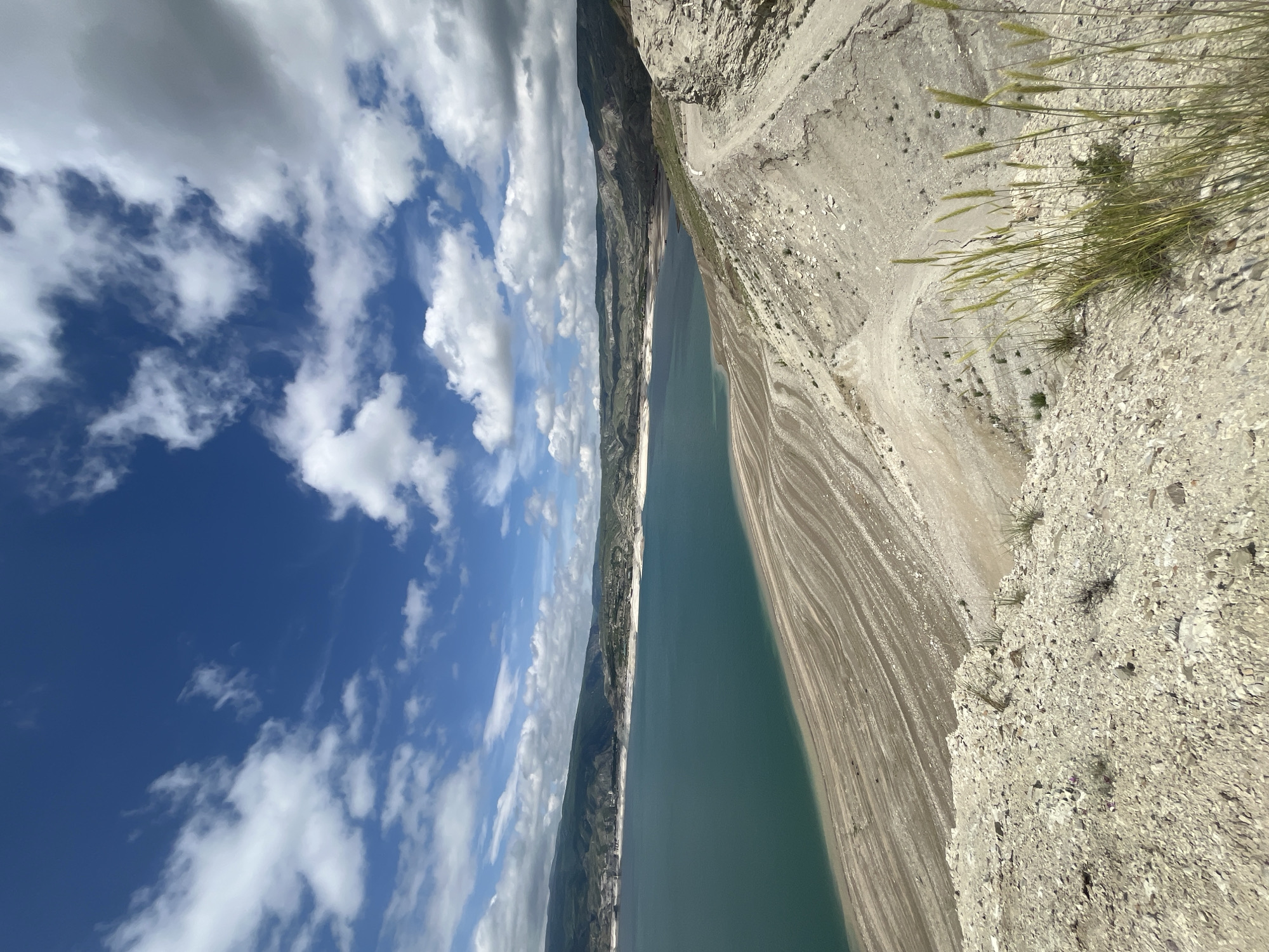 A vertical shot of the Chirkey Reservoir in Chirkey, Russia shows a large body of water bordered by rocky terrain on the right and rolling hills with clouds in the background to the left. The rocky terrain has sparse vegetation, while the hills have more, darker vegetation. The sky is blue with scattered cumulus clouds. The water appears to be a slightly greenish-blue color.