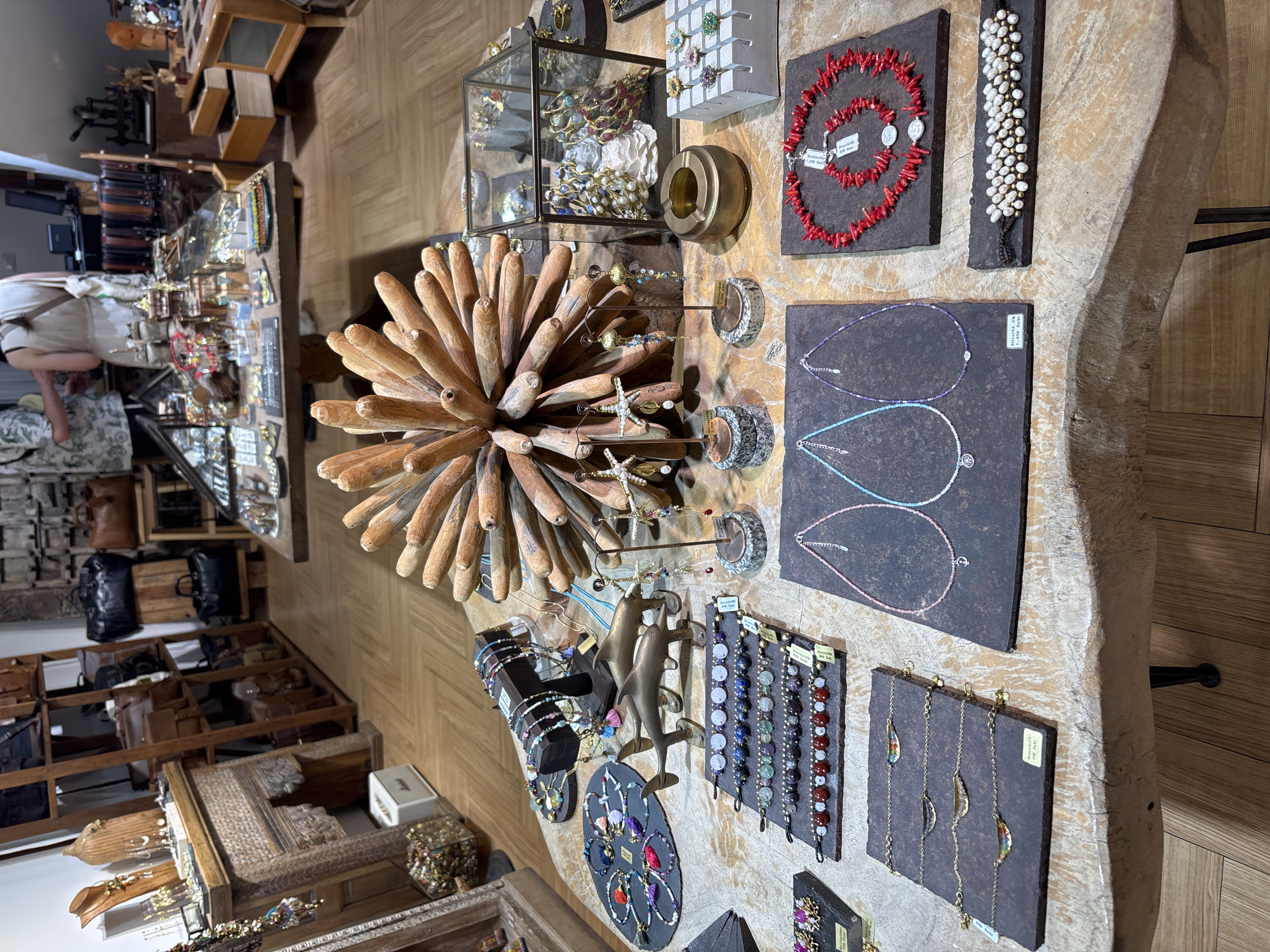 The image depicts a well-lit, indoor retail space, likely a jewelry store in Baan Bo Phut, Thailand, characterized by its wooden flooring and displays. The foreground is dominated by a rustic, carved wooden table laden with various handcrafted jewelry pieces. On the left side, a circular display holds multiple beaded bracelets and necklaces, some adorned with colorful tassels. To the right of this, a dark wooden stand showcases more beaded necklaces. In the center, two bronze dolphin sculptures are positioned on a dark slate base. Several rows of beaded bracelets, featuring a variety of stones and pearls, are neatly arranged on dark slate tiles. Further to the right, a glass display case contains elaborate necklaces and other ornaments, with a round brass ashtray in front of it. A concrete display stand with several rings is also visible. In the lower right quadrant, a vibrant red coral necklace and bracelet set is displayed on a dark slate. Adjacent to it, a pearl and gold bead bracelet is presented on a dark surface. The middle ground shows shelves stocked with leather bags and other merchandise. In the background, two individuals, one wearing a patterned shirt and the other a white top, are interacting near a counter with various retail items. The overall atmosphere is that of a boutique shop offering artisanal jewelry and accessories.