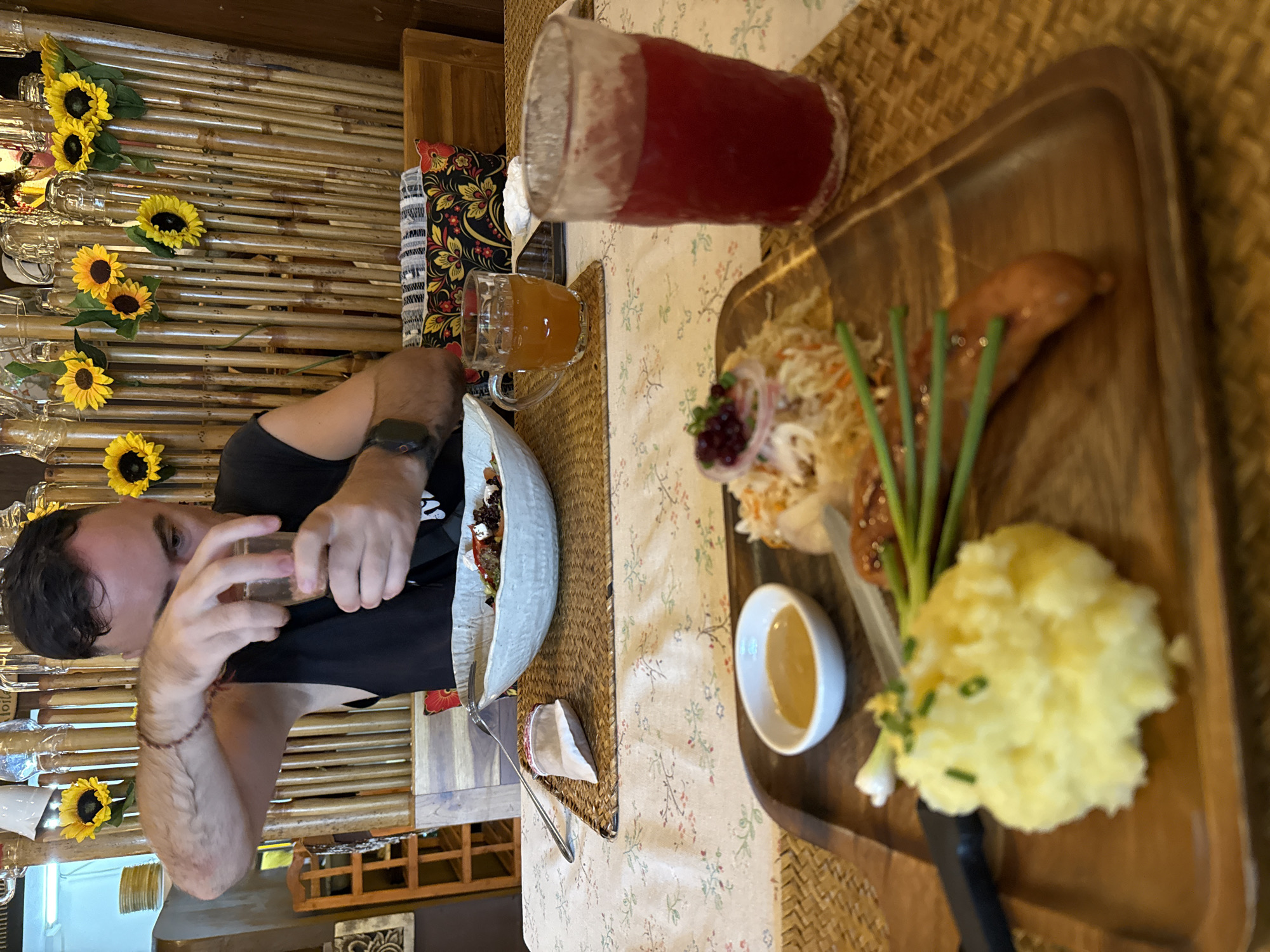 A man seated at a table is holding a glass of liquid, possibly engaging in a toast, in a restaurant setting in Baan Chaweng Noi, Thailand. The table is covered with a floral patterned tablecloth, and is adorned with a wooden serving tray containing a meal that includes sausage, coleslaw, mashed potatoes with chives, and a small dish of sauce. There are also two glasses with beverages: one containing a red liquid and another a brown or orange-colored drink. Other dishware and a napkin are on the table alongside the man. The restaurant decor includes sunflowers decorating a textured wall that appears to be made of bamboo, and decorative drinking glasses along the same wall.