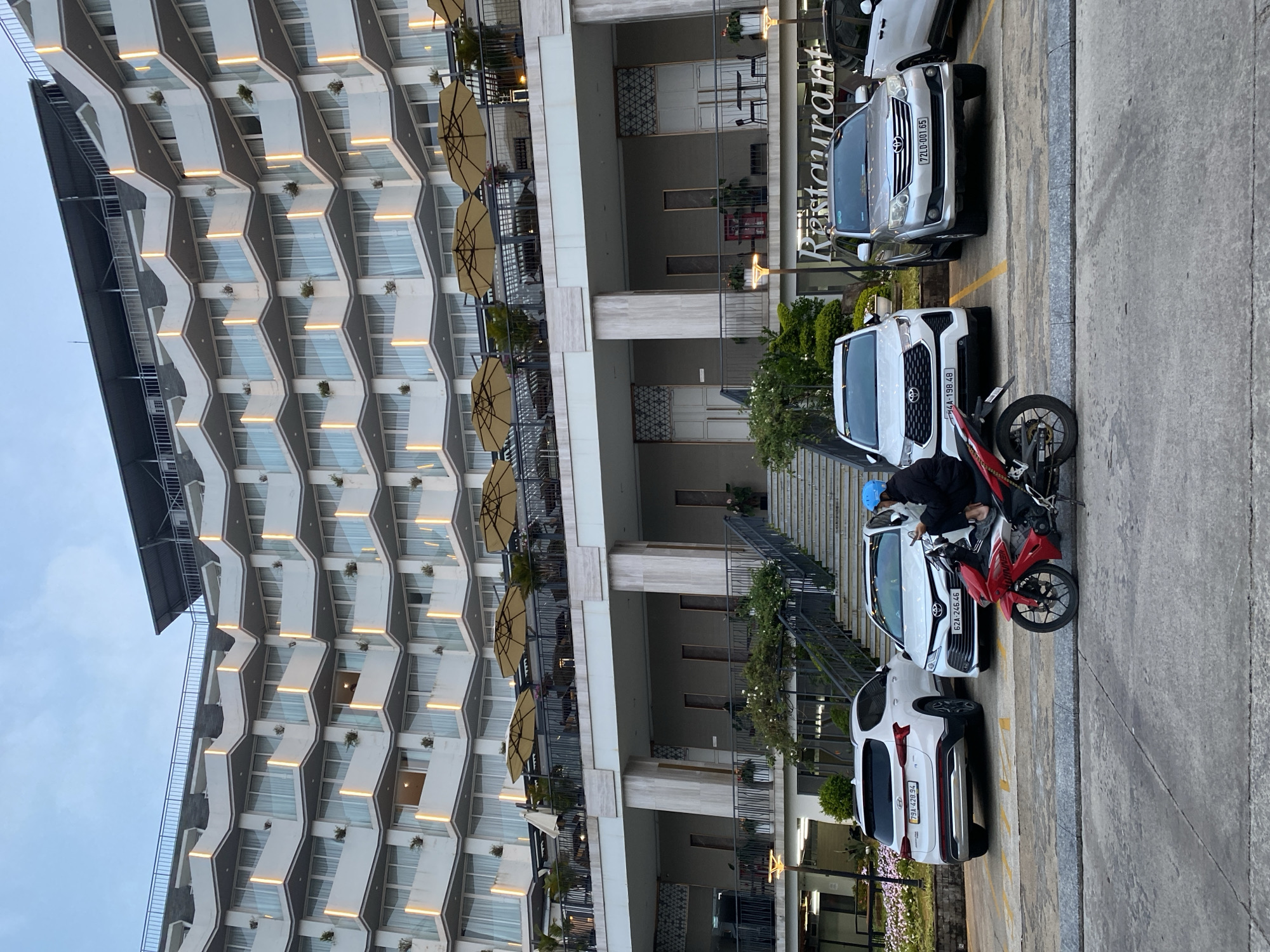 The image captures a scene in Da Lat, Vietnam, in front of a modern building with a distinctive honeycomb facade. The building features multiple balconies with tables and umbrellas. A restaurant sign is visible on the ground floor. The foreground shows a parking area with several cars parked, including a silver SUV (license plate 72LD-001.65) and a white car (license plate 62A-198.48). A red and black motorbike is parked between the white car and another white SUV. A person wearing a blue helmet is near the motorbike, seemingly adjusting something on the bike. The environment suggests an urban setting with a modern architectural style.