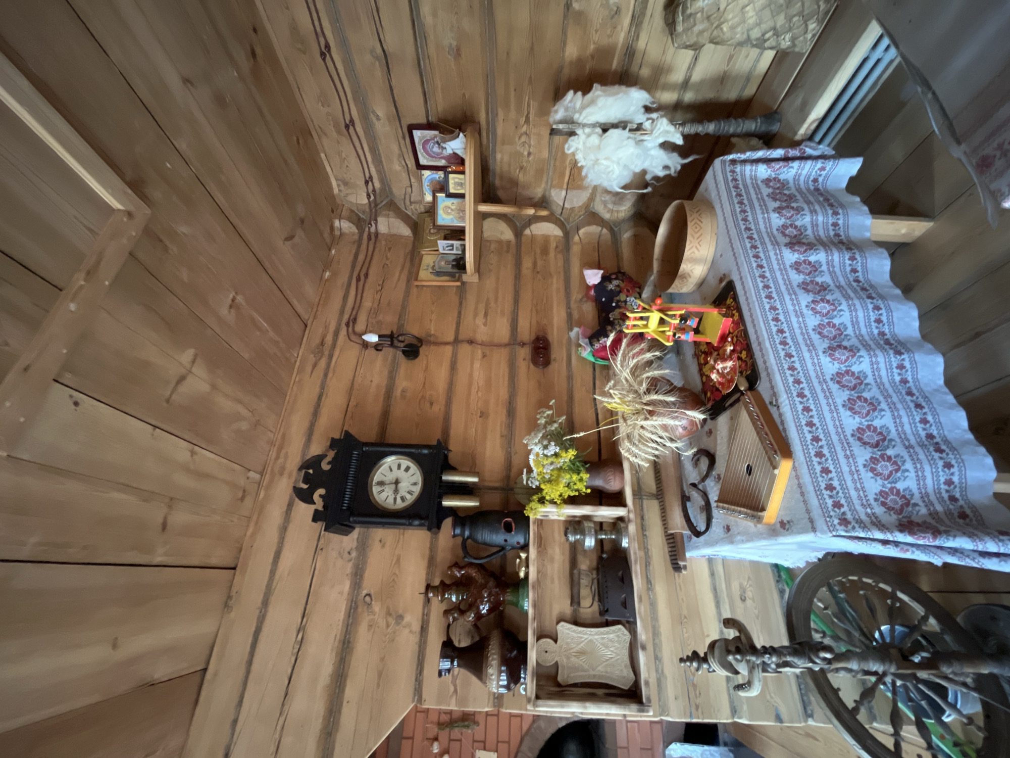 This media file captures a rustic interior scene, likely a traditional Russian dwelling in Vybolovo, Russia, given the context. The walls and ceiling are constructed from wooden planks, creating a warm, textured environment. Various objects are displayed, suggesting a collection of folk art or historical artifacts.
Prominently featured is a black, ornate grandfather clock with a visible clock face. Adjacent to it are shelves holding religious icons, a wooden bowl, and decorative items. Further to the right, a table is draped with a white cloth adorned with a red floral pattern. On this table, a kantele-like musical instrument, a wooden vase with dried wheat stalks, and other small items are arranged. A spinning wheel is visible in the lower right portion of the image, indicating a historical context related to textile production. Scattered throughout the scene are various other decorative and functional objects, including ceramic vessels and what appears to be raw wool. The overall impression is one of a well-preserved, traditional home or museum exhibit, showcasing elements of rural Russian life and culture. There is no indication of people or a specific event taking place. The lighting suggests daytime, and the interior setting is cozy and filled with historical charm.