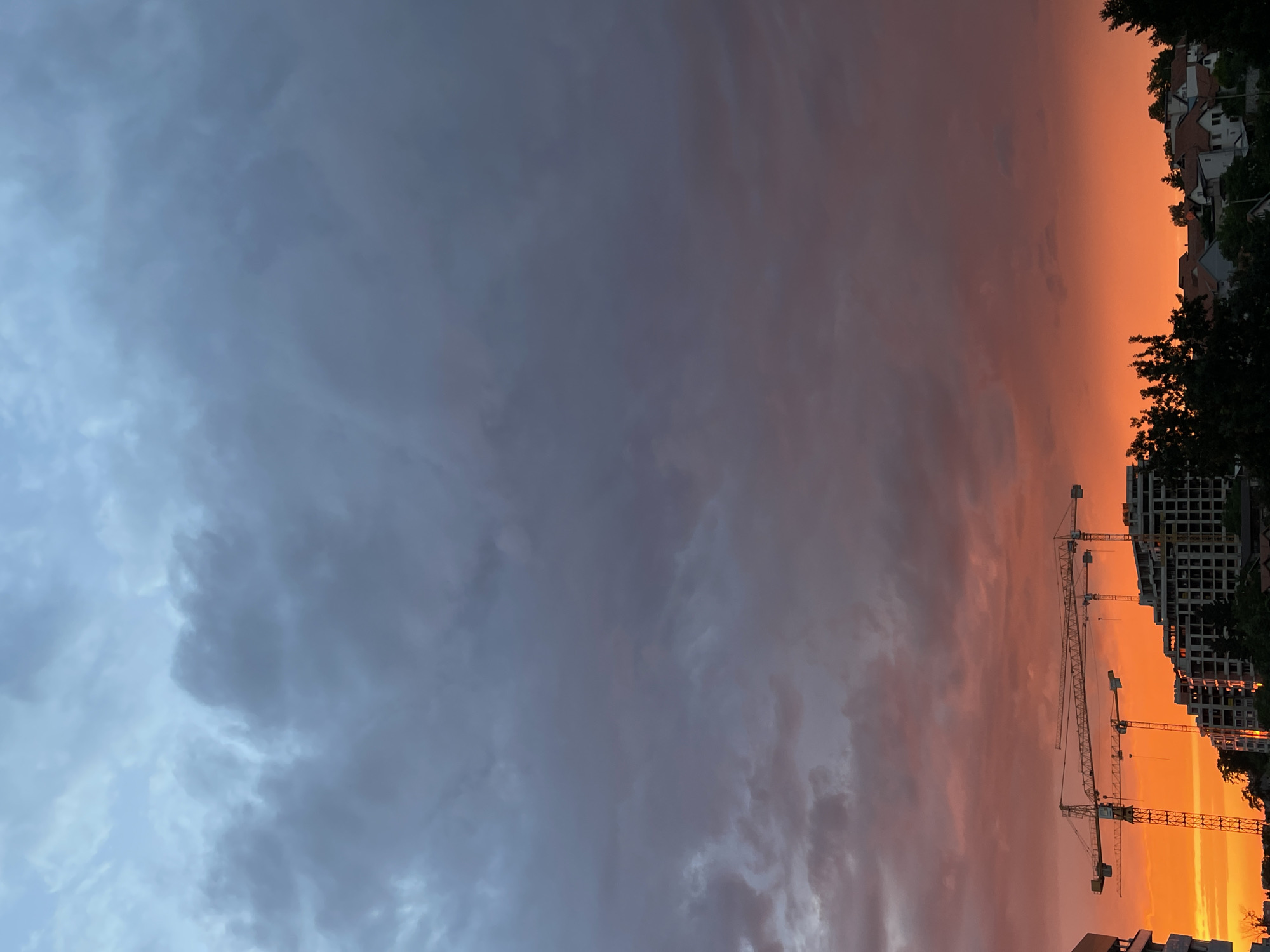 This is a vertical, eye-level shot of a vibrant sunset in the Zvezdara Urban Municipality, Serbia. The sky is a dramatic canvas of color, transitioning from a bright, fiery orange at the horizon to a deep purple and then a muted blue-grey overhead. Dark, heavy clouds dominate the upper portion of the sky, with lighter wisps of clouds catching the sunset's glow lower down.
In the foreground, construction cranes are silhouetted against the intense orange glow. Three large cranes are visible, their latticed structures stark against the sky. Below them, a multi-story building under construction, with visible windows and external structural elements, rises from the ground. To the right and lower, a cluster of residential buildings with red-tiled roofs is nestled among dark green trees, suggesting a developed urban or suburban area. The overall impression is one of ongoing development juxtaposed with the natural spectacle of a dramatic sunset. No people are visible in the frame.