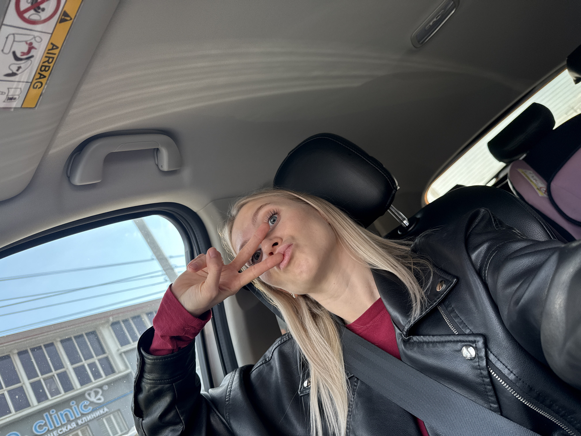 The image is a selfie taken from inside a car. A young woman with long, blonde hair is making a peace sign with her hand and puckering her lips. She is wearing a red shirt and a black leather jacket. A seatbelt is visible across her chest. In the background, through the car window, is a building with the word "clinic" written in English and Russian ("КЛИНИКА"). The building's facade is white with dark window frames. Based on the Cyrillic text and building style, it is likely the photo was taken in Izhevsk, Russia.