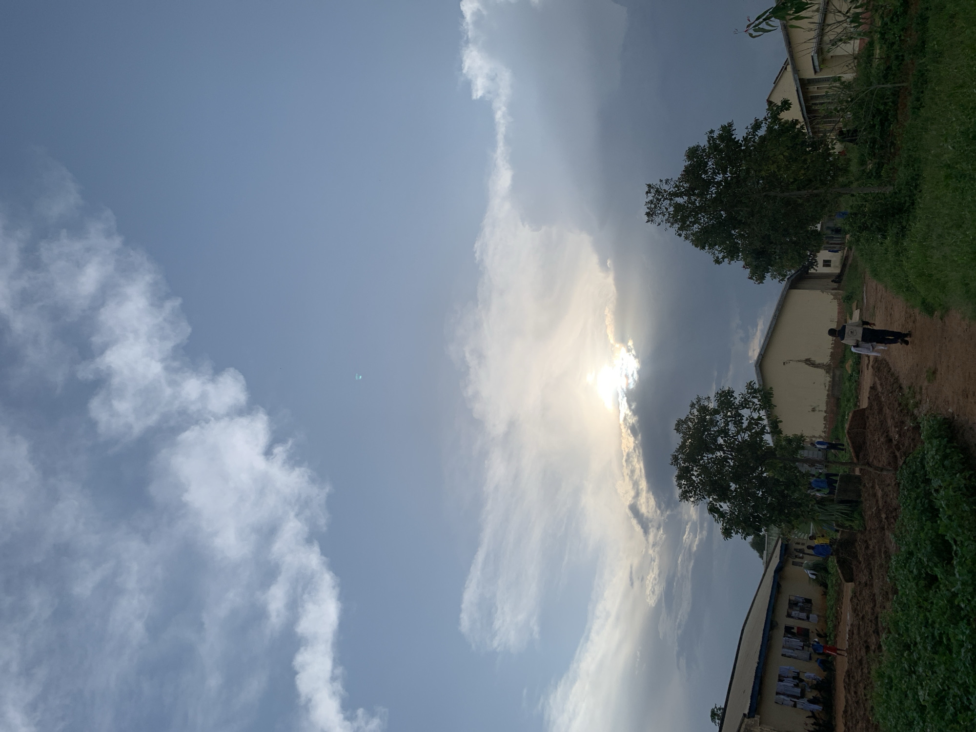 The image depicts an outdoor scene with a bright sky filled with dramatic clouds, some illuminated by the sun, which is partially obscured. The sun's rays are visible breaking through the clouds, suggesting it might be late afternoon or early morning. The scene includes a dirt path running alongside a building, with lush green vegetation on the right side of the path. Two buildings with light-colored exteriors and dark roofs are visible.
Several people are present. One person is walking on the path away from the camera, wearing dark clothing and carrying something in their hands. Another individual, wearing a blue shirt and dark pants, is standing near the first building. Further down the path, a few more people, some in blue attire, are visible near the second building. There are posters or papers displayed on the exterior wall of the second building. A tree with full foliage stands between the two buildings.
The overall atmosphere appears calm, with the natural elements of sky, clouds, and greenery dominating the composition. The location is identified as Kuru Karama, Nigeria. The weather seems to be a mix of sunny and cloudy, creating an interesting interplay of light and shadow.