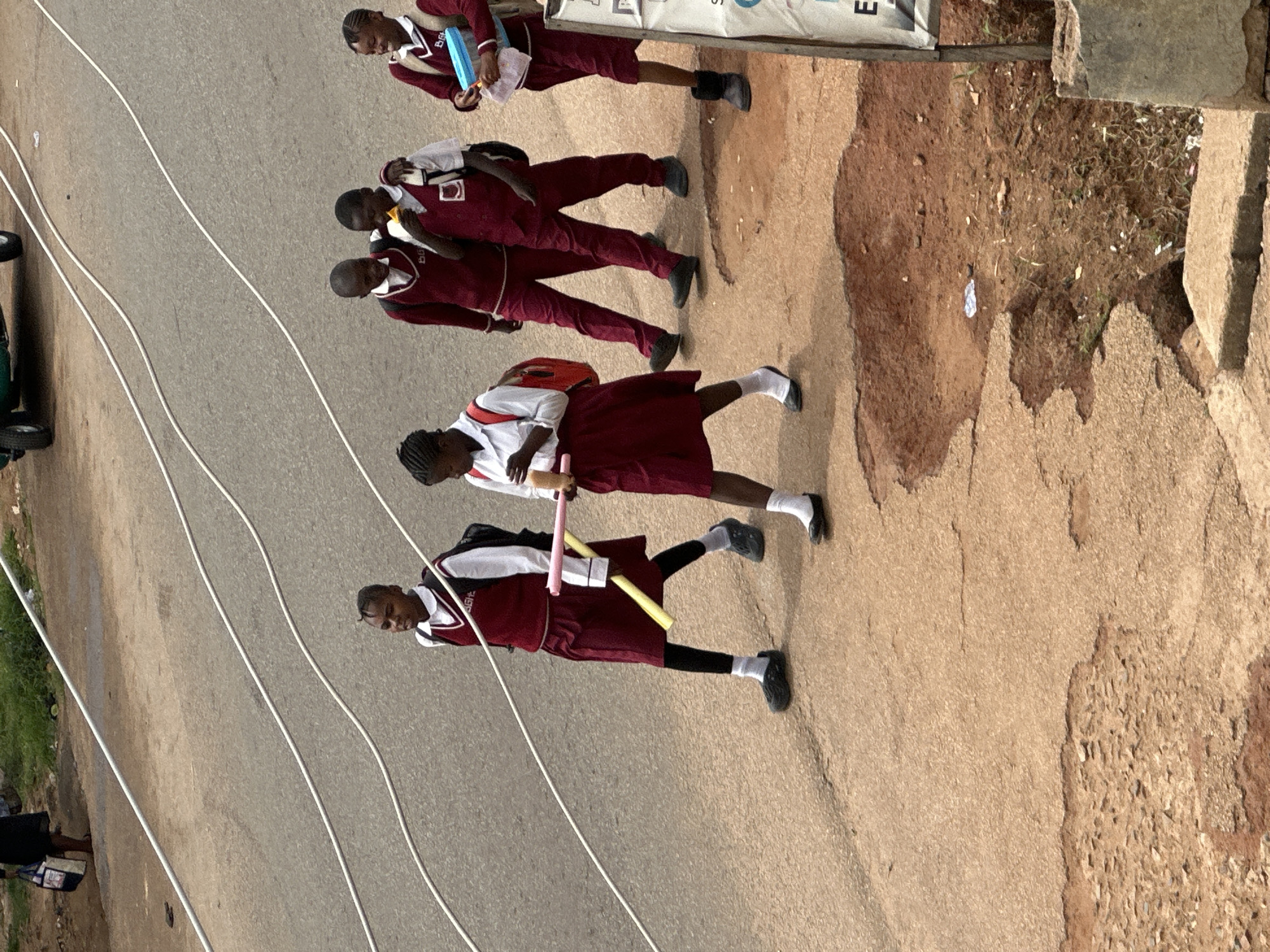 A group of five schoolchildren, dressed in maroon and white school uniforms, walk along a paved road in Jos, Nigeria. The children are walking in a line from left to right. The child on the far left is a girl wearing a maroon vest over a white shirt, a maroon skirt, black leggings, and black shoes with white socks. She is carrying a red backpack and holding a yellow and pink cylindrical object. Next to her are two boys, both wearing maroon vests over white shirts and maroon pants. The boy on the left has his arm around the boy on the right, who is eating something. The fourth child, a girl, is wearing a similar uniform and a red backpack. She is holding a blue container. The fifth child, also a girl in a maroon vest over a white shirt and maroon skirt, is partially visible on the far right. The road appears worn and cracked in places, with a patch of exposed red earth and debris on the right side of the image. Several white cables or pipes run across the road. In the background, a green vehicle is partially visible, along with some grass and a person carrying a bag on the far left.