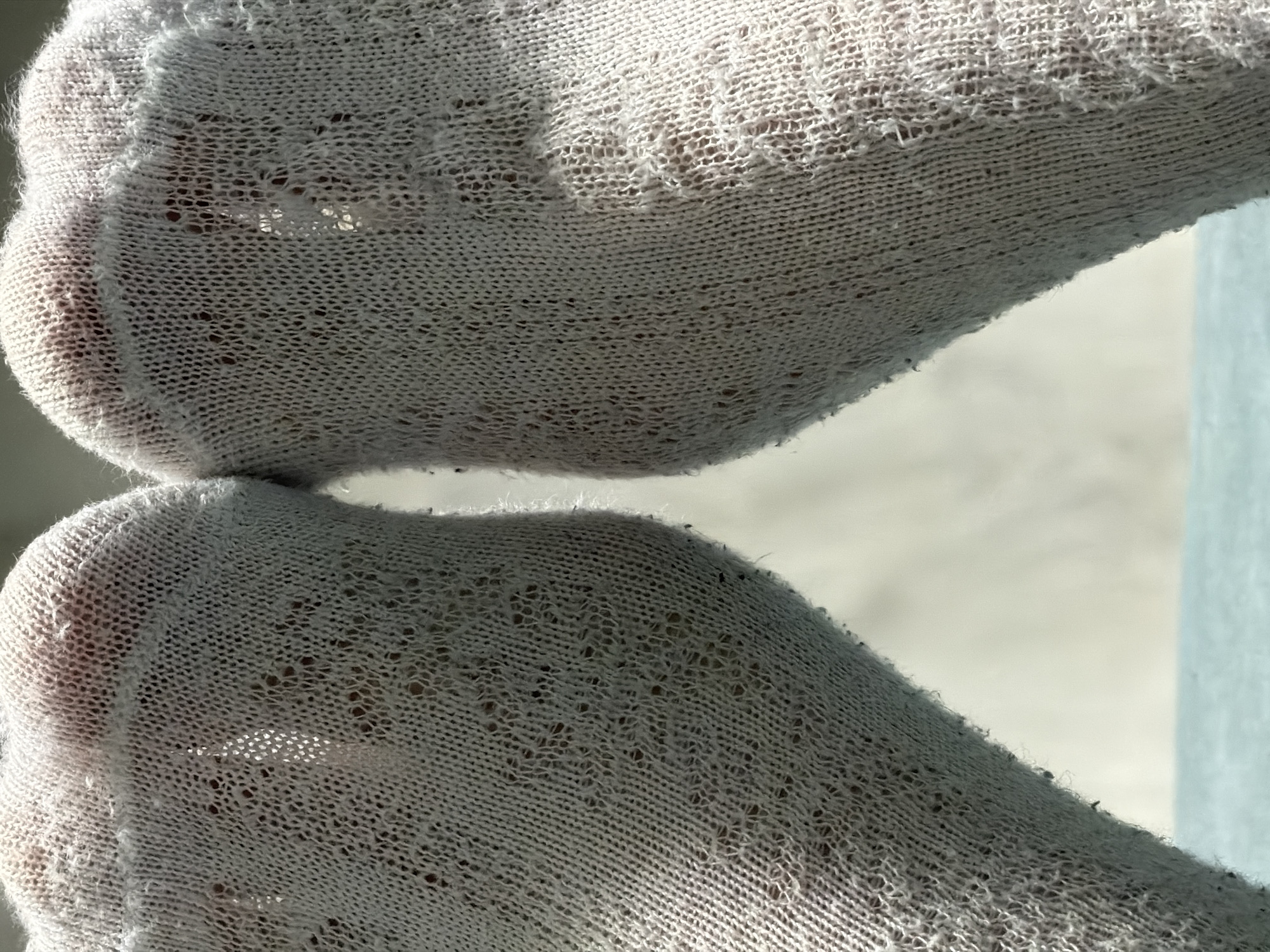 The image is a close-up, low-angle shot of two feet wearing white, sheer, patterned socks. The socks have small, diamond-shaped cutouts and are somewhat translucent, allowing the skin beneath to be visible. Sunlight is streaming in from the left, creating highlights and shadows on the socks and casting a light on the background. The feet are positioned close together, with the toes pointing slightly upwards and towards the viewer. The surface beneath the feet appears to be a light blue or turquoise color, possibly a table or floor. The background is out of focus, showing a lighter, neutral color. There is no indication of a specific location in Naberezhnye Chelny, Russia, or any context beyond the close-up view of the socks and feet.