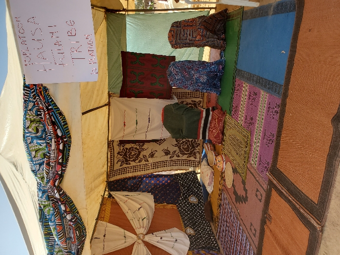 The image depicts the interior of a white canopy tent in Bar Gada, Nigeria, serving as an exhibition or display area. A white sign handwritten with "10 PLATOON HAUSA FULANI TRIBE 10 PLATOON" hangs from the tent's internal frame. The tent walls are draped with various patterned textiles, including white embroidered fabric, dark blue geometric prints, beige floral patterns, and dark red/green designs. A colorful patterned fabric valance adorns the front upper opening. The floor is covered by an arrangement of interlocking rectangular woven mats in orange, pink, blue, green, and brown hues. Positioned on the floor towards the rear are a light-colored, possibly embroidered cushion, and two circular woven caps. Three upright figures, resembling mannequins, are draped in distinct patterned fabrics: one in dark green with red and black horizontal stripes, another in blue patterned textile, and the third in brown geometric patterned fabric. No individuals are present in the scene.