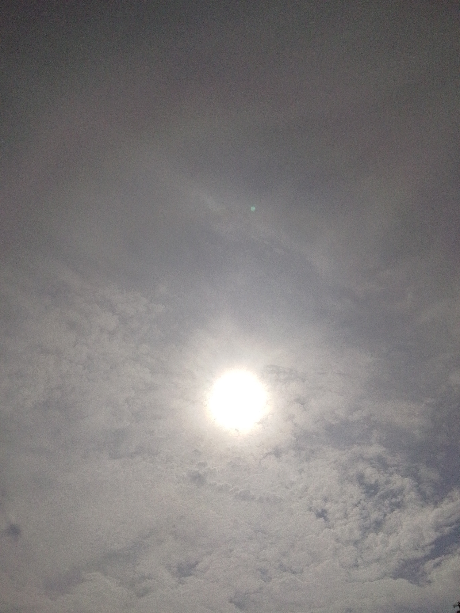 The image shows a cloudy sky with the sun partially obscured by clouds. The sun appears as a bright, white disc in the center of the image, with a slight green lens flare visible to the upper left of the sun. The clouds are white and puffy, with some darker areas suggesting depth and texture. The overall impression is one of a bright but overcast day. The location is the city of Serti, Nigeria.