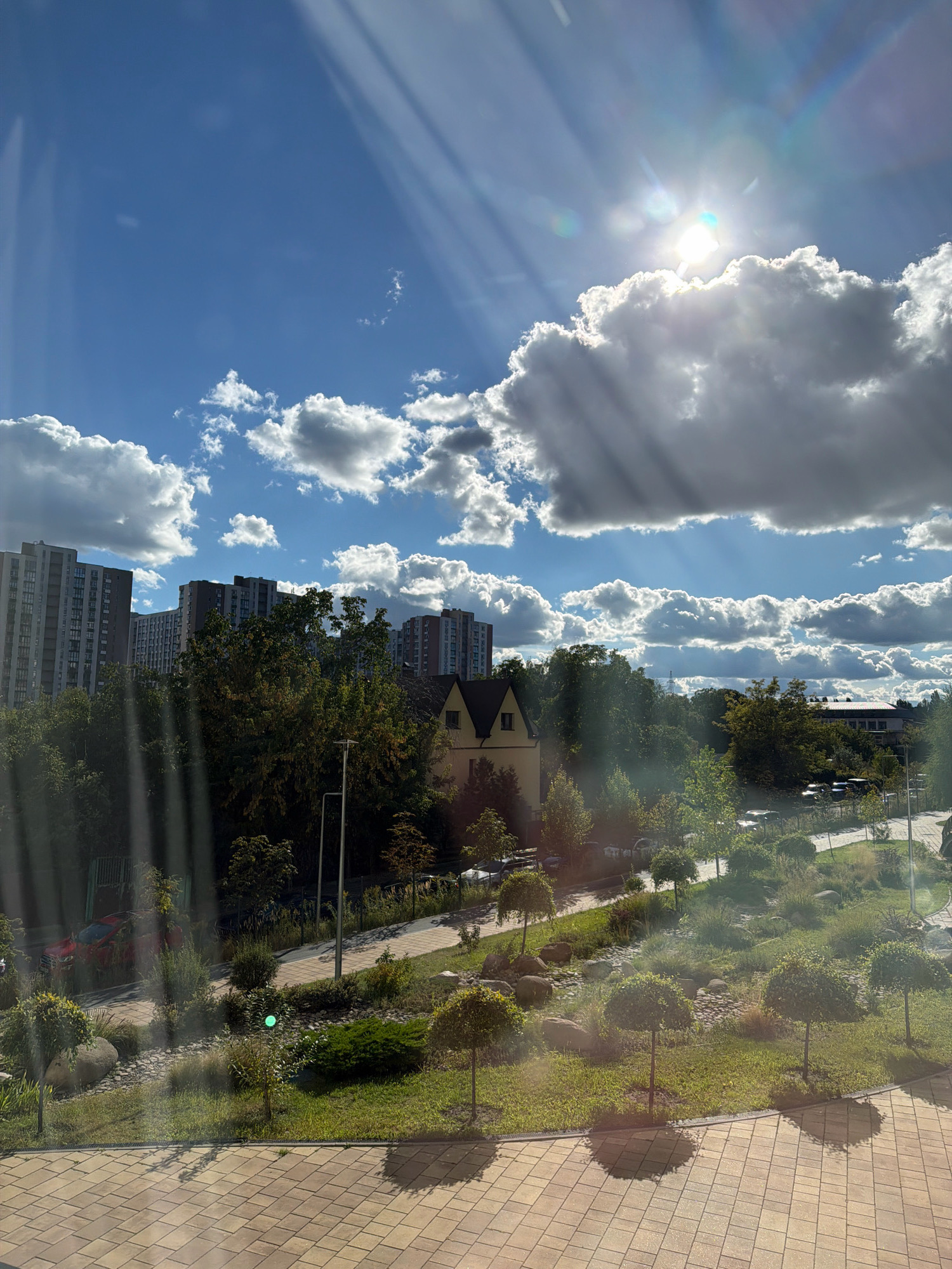 This is an eye-level, medium shot of a city park area in Kyiv, Ukraine, with a bright sun and clouds visible in the sky. The image captures a residential area with tall apartment buildings in the background, and a park with manicured trees, grassy areas, and pathways in the foreground. The sun's rays are streaking through the clouds, creating a dramatic lighting effect. There are no people or animals visible in the scene. The overall atmosphere is serene and peaceful, with the sunlit park area contrasting with the urban backdrop.