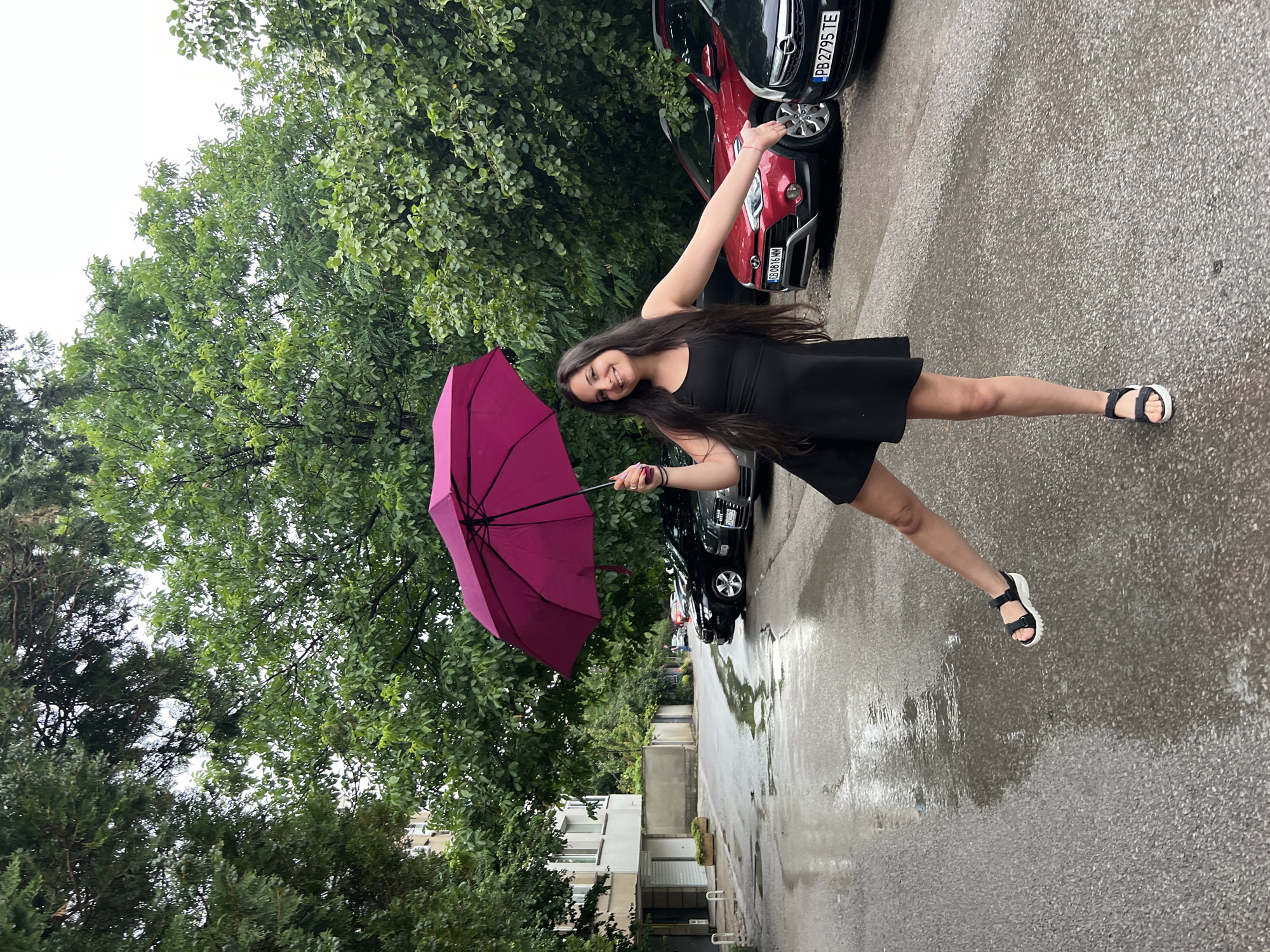 A young woman with long, dark hair stands outdoors holding a magenta umbrella. She is wearing a black dress and black sandals with white soles. Her right arm is extended upwards and to the side, while her left hand holds the umbrella. She is smiling at the camera. The ground is wet and reflects the surroundings, indicating it has recently rained or is raining. Several cars are parked in the background, with license plates visible. The scene is set against a backdrop of lush green trees and a building. The weather appears overcast, suggesting it might be a cloudy day or the aftermath of rain. The location is identified as Sofia, Bulgaria. The license plates on the cars show text, including "PB 2795 TE" and "CB 0816 MM". The woman appears joyful and playful.