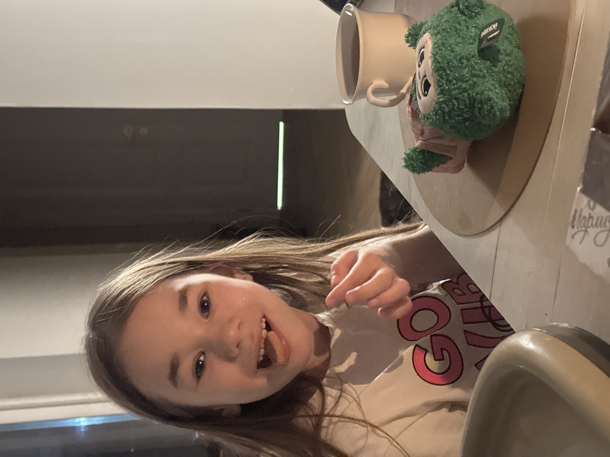 A young girl sits at a table with a mug, a green plush toy, and another object partially visible on the right. The girl has brown hair, fair skin, and is wearing a light-colored shirt with pink letters. She is smiling and appears to have a food item on her tongue. The background includes a door or opening in a dimly lit room. This photo does not appear to be taken in the city of Kharnavy, Russia and there is no evidence in the image to suggest it was taken there.