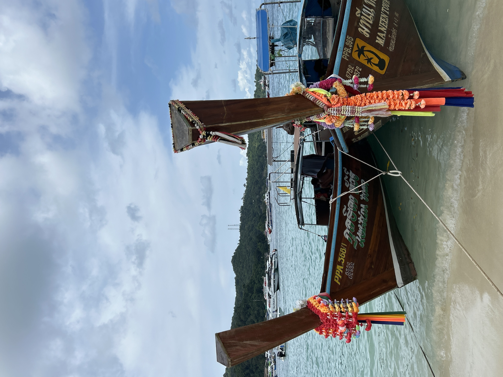 This is a close-up, eye-level, vertical shot of two traditional Thai longtail boats docked in shallow, clear turquoise water at the shore of Ao Nang, Thailand. The boats are made of dark wood and are decorated with colorful leis of flowers and streamers. The boat on the left has the registration number "PPA.368/1" and some Thai script. The boat on the right has the registration number "PPB.395" and also Thai script with a silhouette of a palm tree. The foreground shows the sandy beach and the water's edge with ropes tethering the boats. In the background, there are more boats anchored in the bay, a lush green, tree-covered island, and a cloudy sky with patches of blue. The overall impression is a typical tourist scene in a Thai coastal town.
