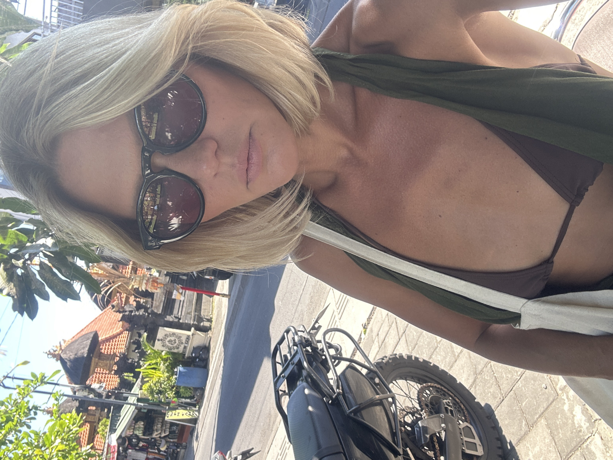 A woman with blonde, shoulder-length hair and sunglasses is taking a selfie outdoors on a sunny day. She is wearing a brown bikini top and a green garment draped over her shoulders. A white canvas bag is slung over her shoulder. To her left, a black motorcycle is parked on a tiled sidewalk. In the background, traditional Balinese buildings with red tile roofs and intricate stone carvings line a street. The scene appears to be in North Kuta, Indonesia.