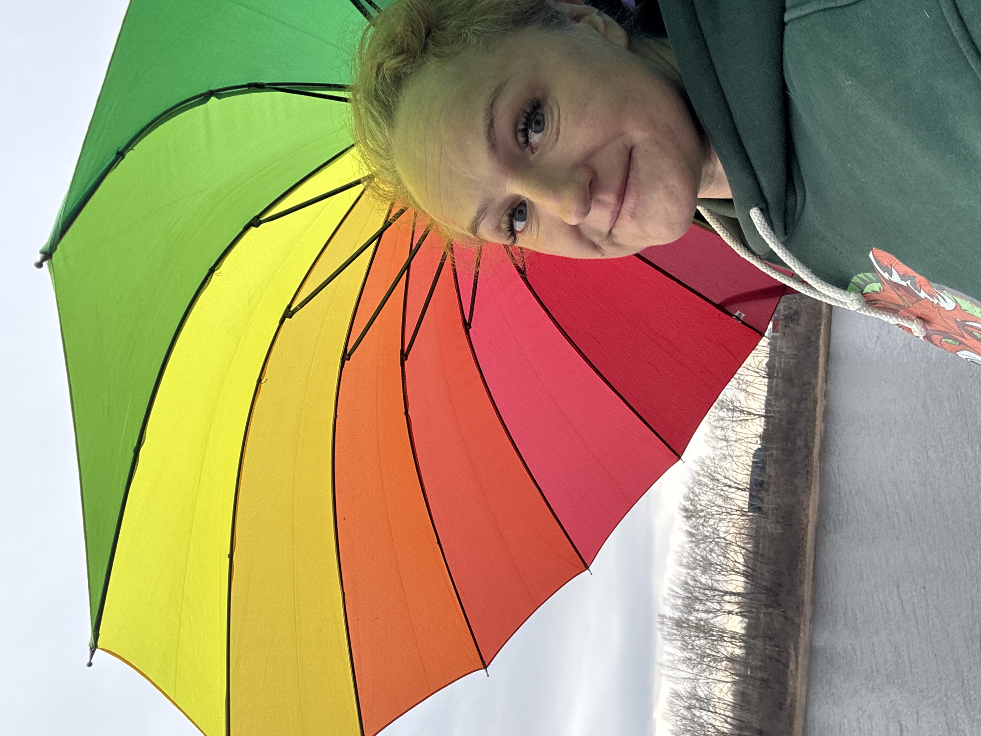 The image shows a fair-skinned woman holding a rainbow-colored umbrella. She is wearing a dark green hooded sweatshirt. Behind the woman and umbrella, there is a body of water, with trees lining the edge. The image was taken outdoors in Chaikovsky, Russia.