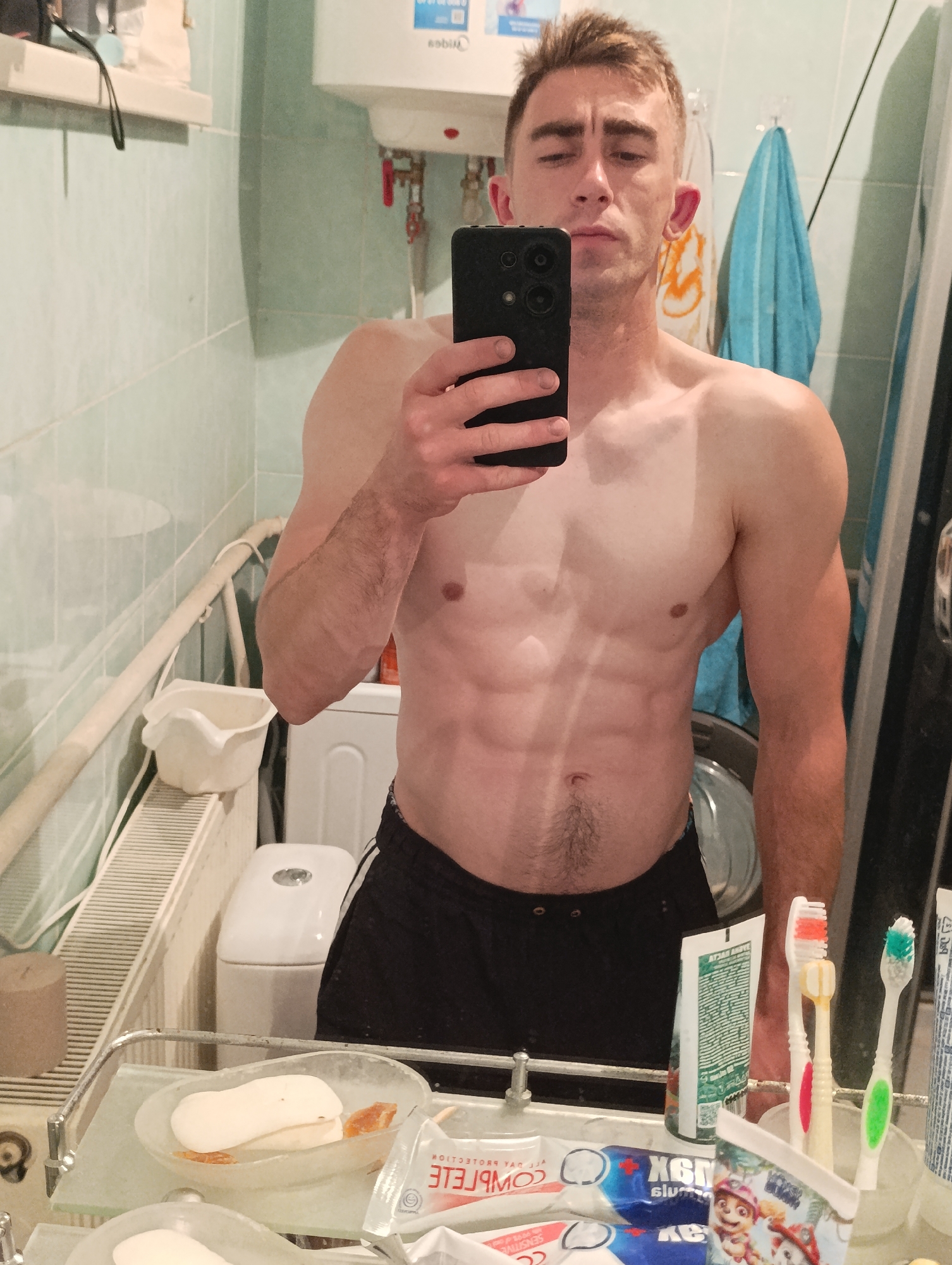 The image is a full-body mirror selfie taken in a bathroom. A man with visible abdominal muscles and short brown hair is holding a smartphone to take the picture. He is wearing black shorts. In the background, there is a white toilet, a sink with a soap dish, toothbrushes, and a water heater. The bathroom has light green tiled walls.