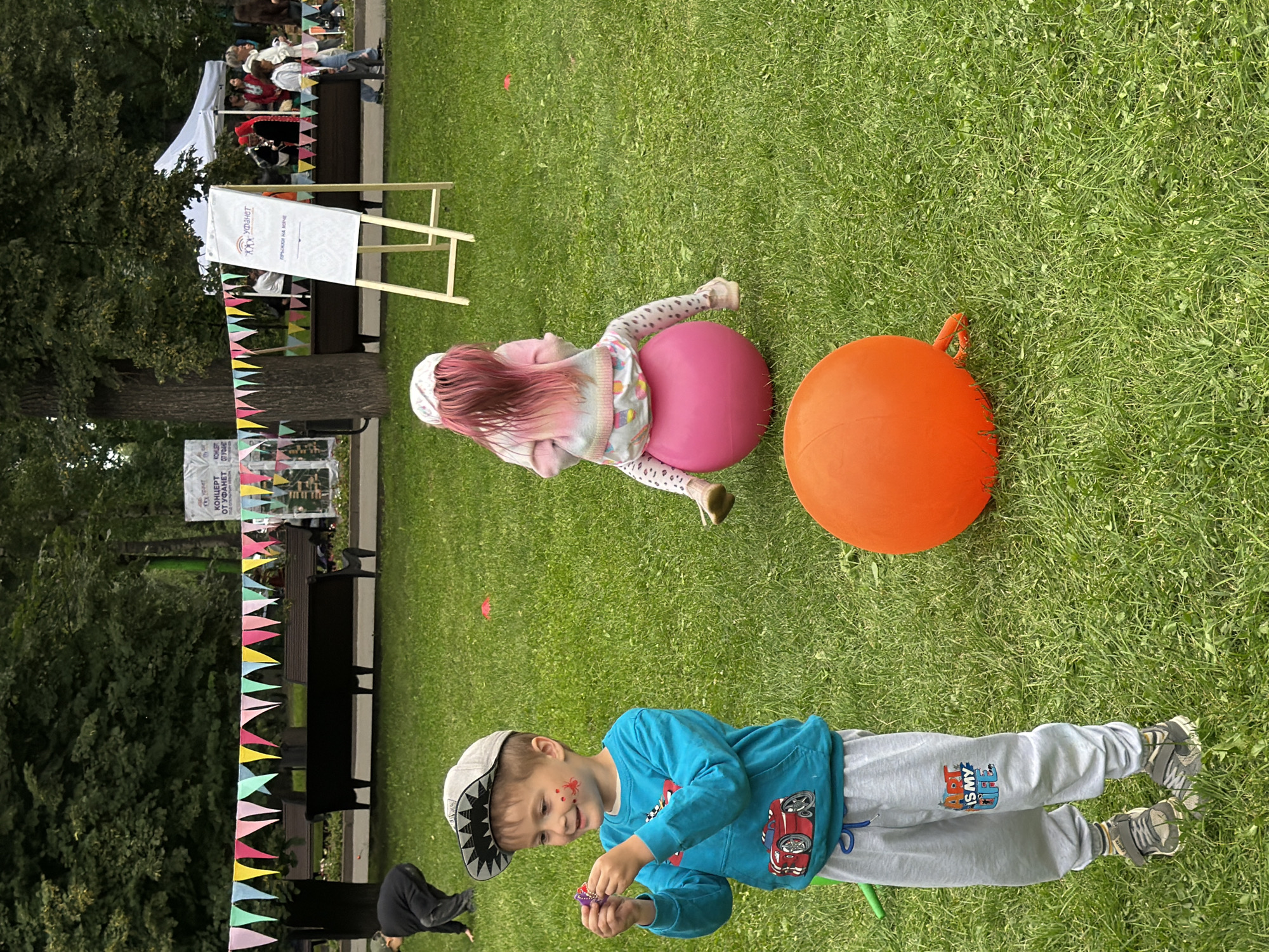 This is a horizontal, eye-level, full shot of an outdoor event on a grassy lawn. The event appears to be a family-friendly festival or gathering, possibly for children, in Kazan, Russia.
The main subjects are two children. In the foreground, a young boy wearing a grey baseball cap with a shark design, a blue long-sleeved shirt with a car graphic, and grey sweatpants is holding a small, purple, bubble-blowing toy. He has red face paint on his cheek, resembling a spider. Behind him, a younger child, possibly a girl, with pink hair, is lying on their back on a pink exercise ball. They are wearing a white hat, a pastel-colored striped sweater, and patterned leggings.
Several large, round, inflatable balls are present: a pink one under the child on the ground and a large orange one to the right of the frame. The setting is a well-maintained lawn with trees visible in the background. A white tent and some wooden benches are also in the background, along with a sign on an easel with Cyrillic text. A string of colorful triangular flags decorates the area.
The lighting suggests it is daytime, likely sunny, given the shadows cast. The overall mood seems cheerful and playful, indicated by the children's activities and the festive decorations.
Visible text includes Cyrillic on the sign in the background, which appears to say "ГОРОД ОТ УДАЧЕТ" and likely relates to the event name or sponsor.