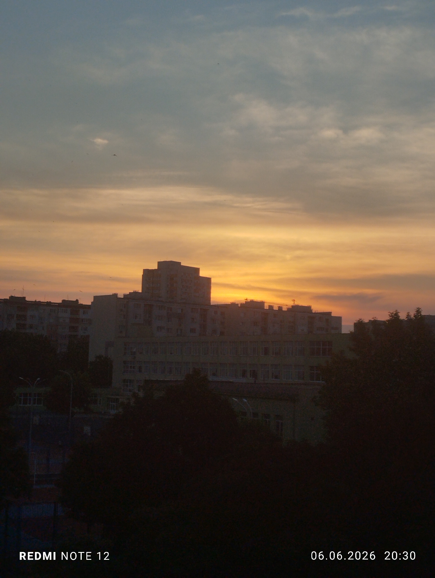 An outdoor photograph captures an urban sunset scene in the city of Burgas, Bulgaria, at 20:30 on June 6, 2026. The sky displays a gradient, transitioning from a light blue-grey at the top to warm shades of orange and yellow near the horizon, with scattered clouds present throughout. Multiple multi-story buildings, characteristic of an urban environment, occupy the midground, appearing as dark silhouettes or partially illuminated against the setting sun. A prominent central building features a stepped roofline. A yellow construction crane is faintly visible atop a building on the left side of the midground. Several small, dark avian forms are discernible in flight against the sky. The foreground consists of dark, dense vegetation and trees, obscuring the ground level, with a partially visible streetlamp on the left. No individuals are present. The image metadata indicates capture by a REDMI NOTE 12 device.