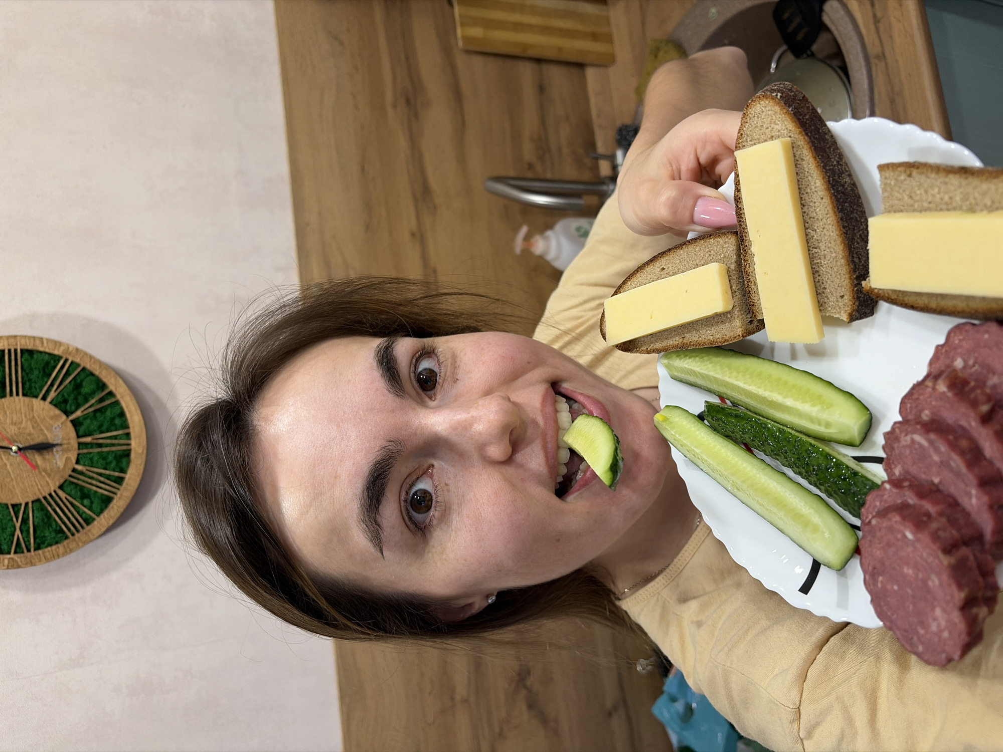 A woman indoors holds a white plate with rye bread topped with cheese, cucumbers, and sliced sausage. She has a cucumber slice in her mouth. A clock is on the wall. A kitchen sink and countertop are in the background. The location is Izhevsk, Russia.