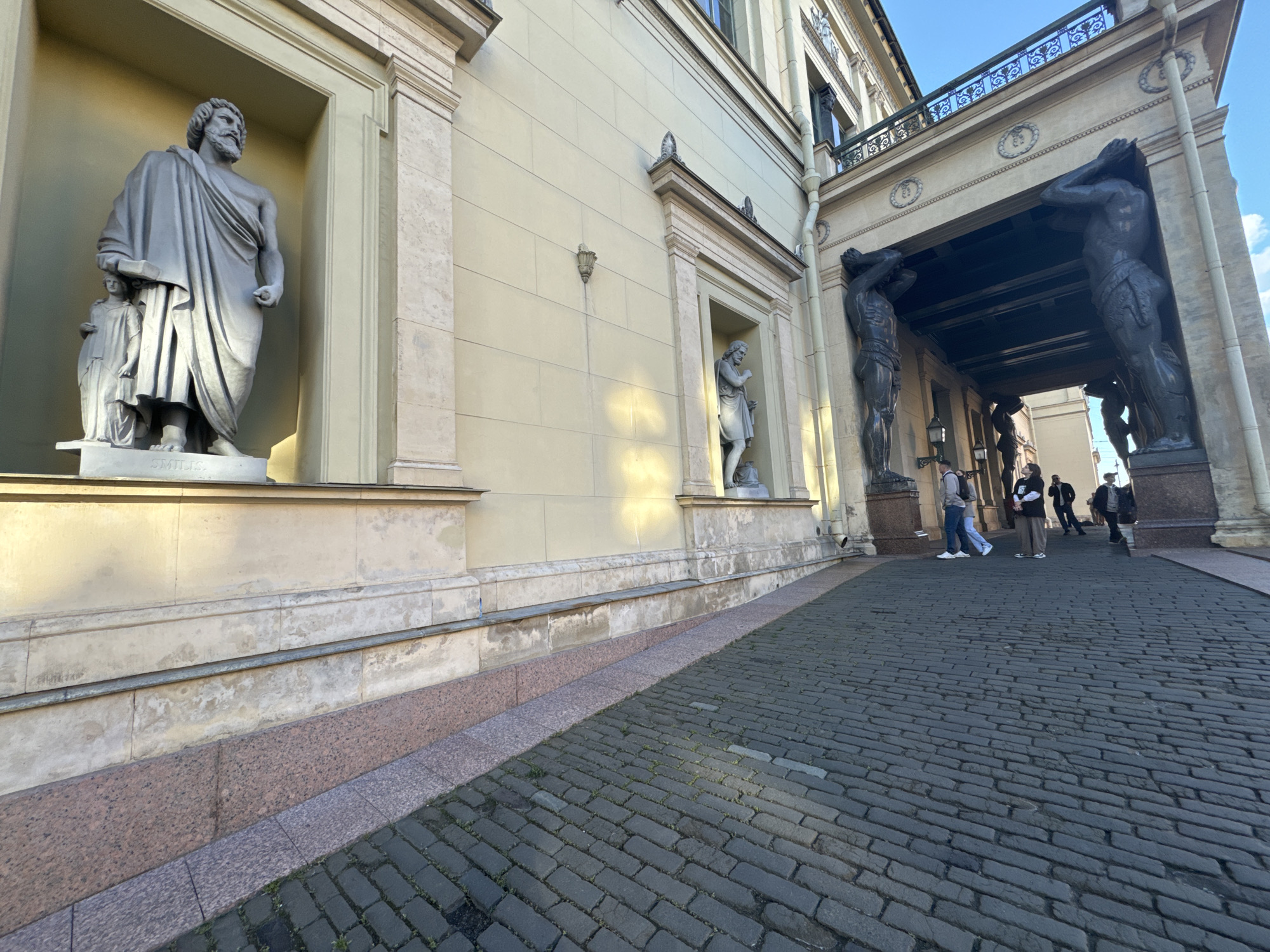 The image shows a street scene in front of a building in Saint Petersburg, Russia. The building has a light-colored facade with classical architectural elements, including statues and columns. In the foreground, a cobblestone pavement leads towards an arched entryway supported by large statues of men, known as Atlantes. Several people are walking on the pavement and near the entryway, some appearing to be tourists. The scene is illuminated by sunlight, casting shadows on the ground and the building. The overall impression is one of a historical and cultural landmark in the city.