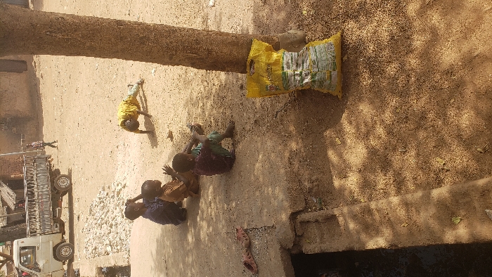 The image depicts an outdoor scene in Kaduna South, Nigeria, under direct sunlight, featuring an unpaved dirt ground. In the immediate foreground on the right, a thick brown tree trunk stands, with a crumpled yellow sack, bearing green and white markings, resting against its base. Adjacent to the tree, a dark-skinned child in a yellow shirt and blue pants is positioned prone on the ground, supported by hands and knees, facing left. Three other dark-skinned children are seated on the dirt ground to the left of the prone child: one in a dark blue garment with hands near their head, one in a light brown top with hands clasped, and one in a dark red top and green pants holding a dark rectangular object. All three seated children face towards the right. To the lower left, a pair of red plastic sandals lies on a concrete slab that partially covers an open dark drain. In the background, a light-colored pickup truck with a utility bed is parked on the left, near a pile of light-colored rubble. Further back, structures with light brown walls are visible. An individual in a plaid shirt and dark pants walks from right to left across the distant background.