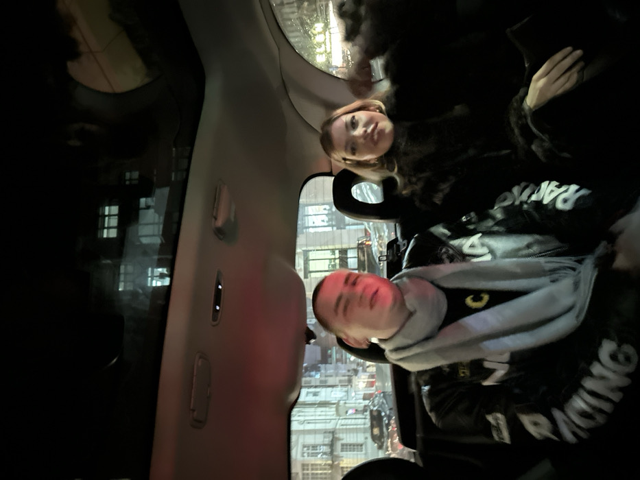 Two people sit in the back of a car. One wears a black leather jacket with "RACING" printed on it, and a gray scarf. The other wears a dark fur jacket. They are visible against a cityscape seen through the car windows. The interior of the car is visible.