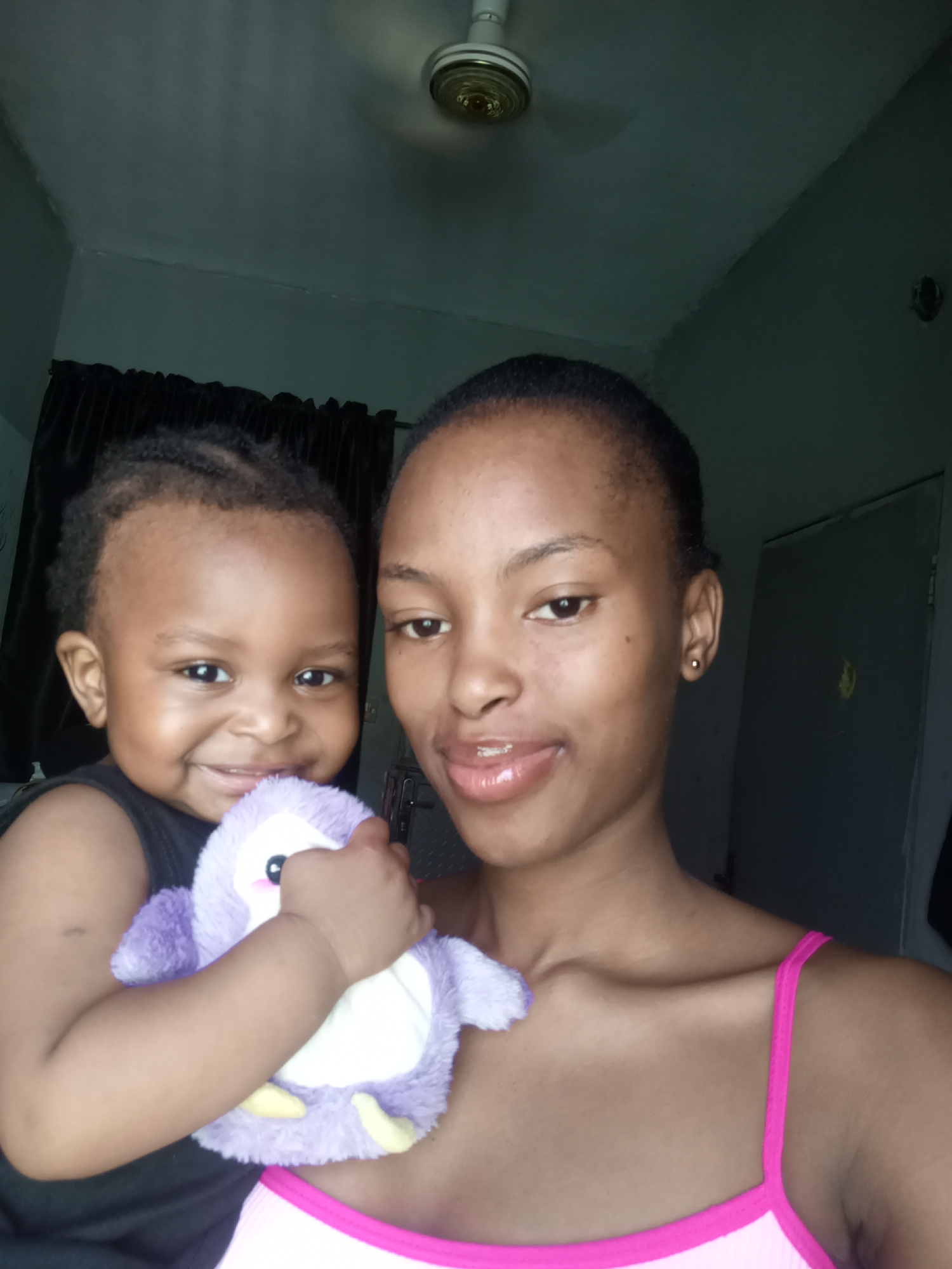A selfie depicts a young woman holding a toddler with a stuffed purple penguin toy. The woman has dark hair pulled back, wearing a pink tank top. The toddler has short, braided hair and wears a black tank top. The background includes a ceiling fan, dark curtains, and a closed gray door. This image was likely taken indoors. There is no direct evidence of the location being in the city of Keffi, Nigeria.