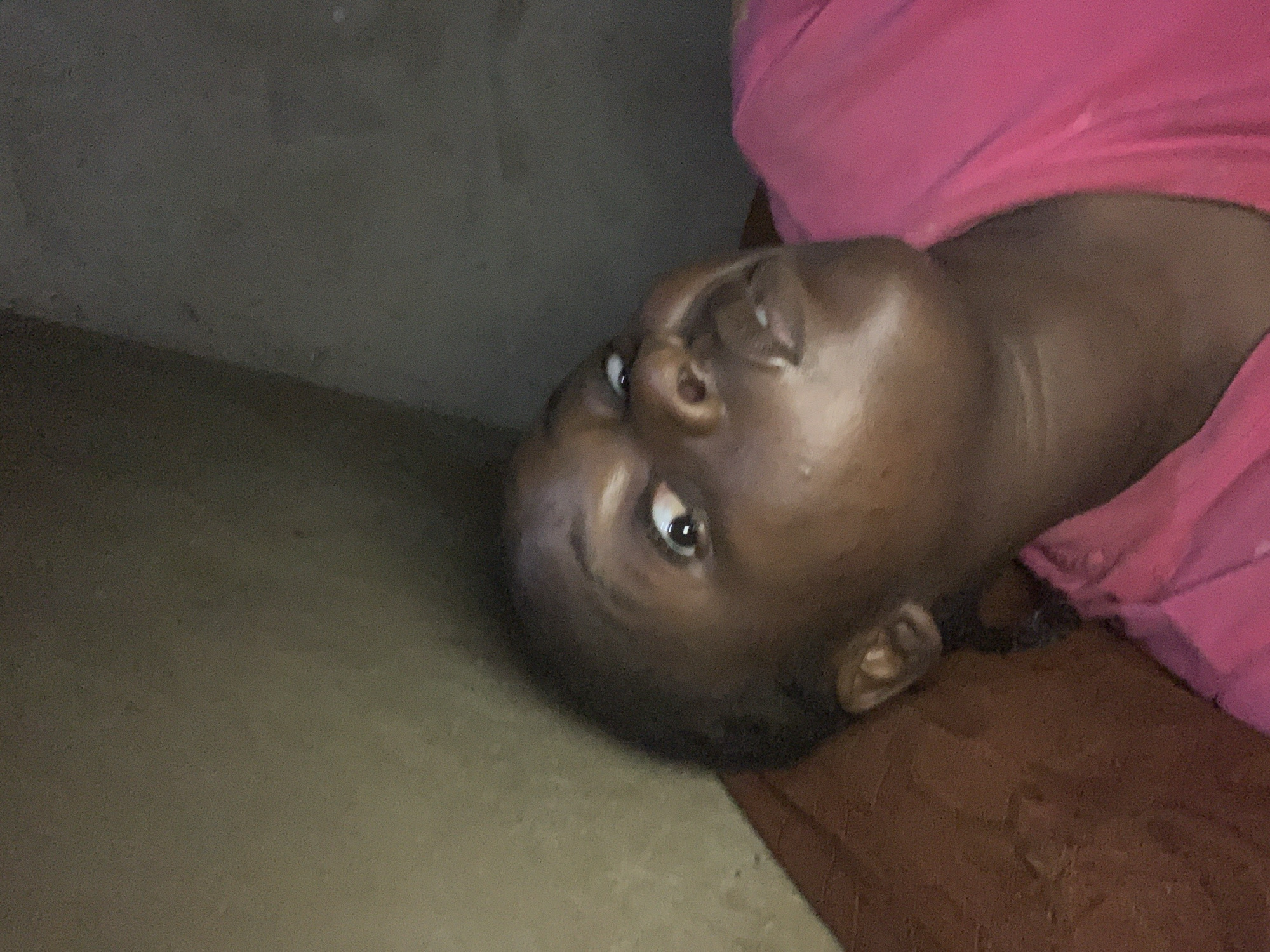 A close-up photograph shows a dark-skinned person lying on a surface, possibly a bed or couch, with their head tilted towards the left of the frame. They are wearing a pink shirt. The background includes a beige wall and brown surface. The location is not discernible as Agban, Nigeria.
