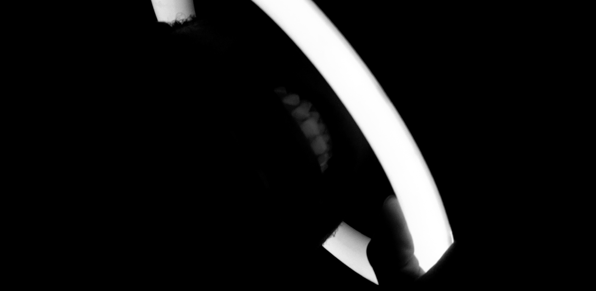 This black and white, high-contrast image presents an abstract close-up, dominated by a bright, curved light source that runs diagonally from the top to the bottom right of the frame. The light appears uniform and intense, suggesting an artificial light source. To the left of this prominent light, a shadowy form reveals several small, lighter, rounded shapes, possibly indicating beaded details on clothing, jewelry, or an object. At the bottom right, a darker, finger-like shape intrudes into the frame, seemingly interacting with or holding the light source. The background is predominantly black, emphasizing the illuminated elements. The scene appears to be taking place in low light conditions, such as at night or in a dark interior. No text is visible.
