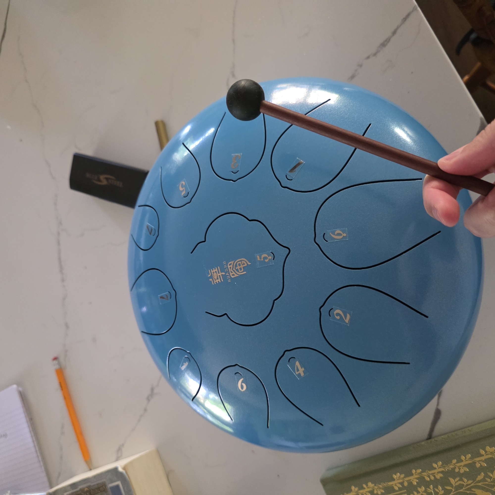 A hand is holding a drumstick with a black rubber ball on the end, poised to strike a sky-blue tongue drum. The drum is positioned on a white speckled countertop, with a marble-like pattern. Several other objects are scattered on the countertop: a thin orange pencil with a red eraser, a small black object with the word "SOL Steel" and an "S" logo on it, a brass rod, and two books. One book on the left is partially visible and appears to be old and bound in green with gold embellishments. Another book on the upper left is open, revealing lines of text. The tongue drum itself has several cut-out sections, each marked with a number or Chinese characters. The numbers 1 through 6 are visible, with the characters "華 樂" and the number "5" in the center. The scene is indoors, suggesting a domestic or studio setting. There is no indication of the city of Portland, United States of America in the image.