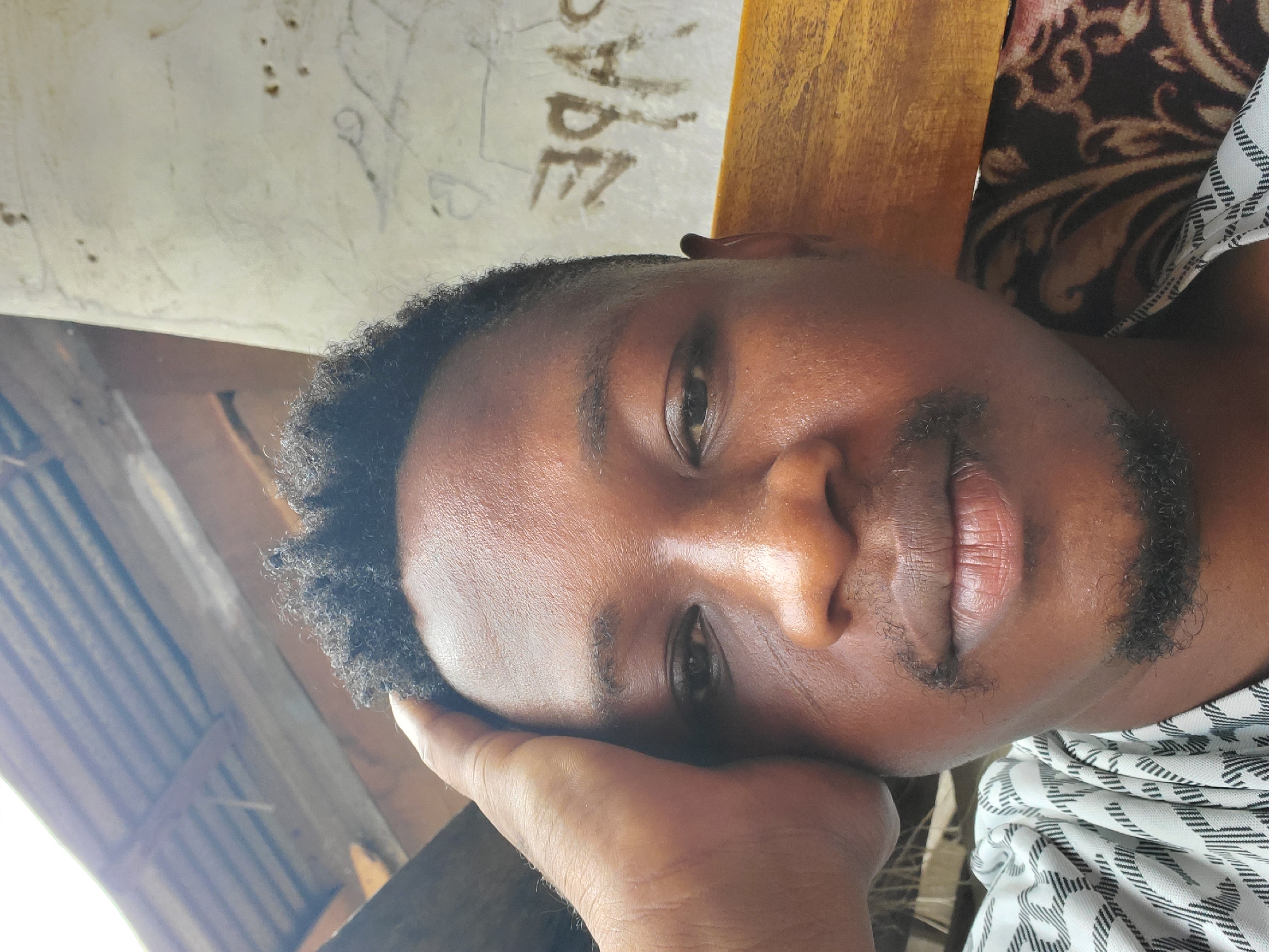 A close-up portrait shows a Black man resting his face on his hand. He has short, dark, curly hair, brown eyes, and a short, dark beard and mustache. He wears a white and black patterned shirt. The background includes a wooden surface, possibly a chair or table, and a patterned fabric. In the upper left, a corrugated metal roof structure is visible. Text is scrawled on the wall. The location is Mararaba, Nigeria.