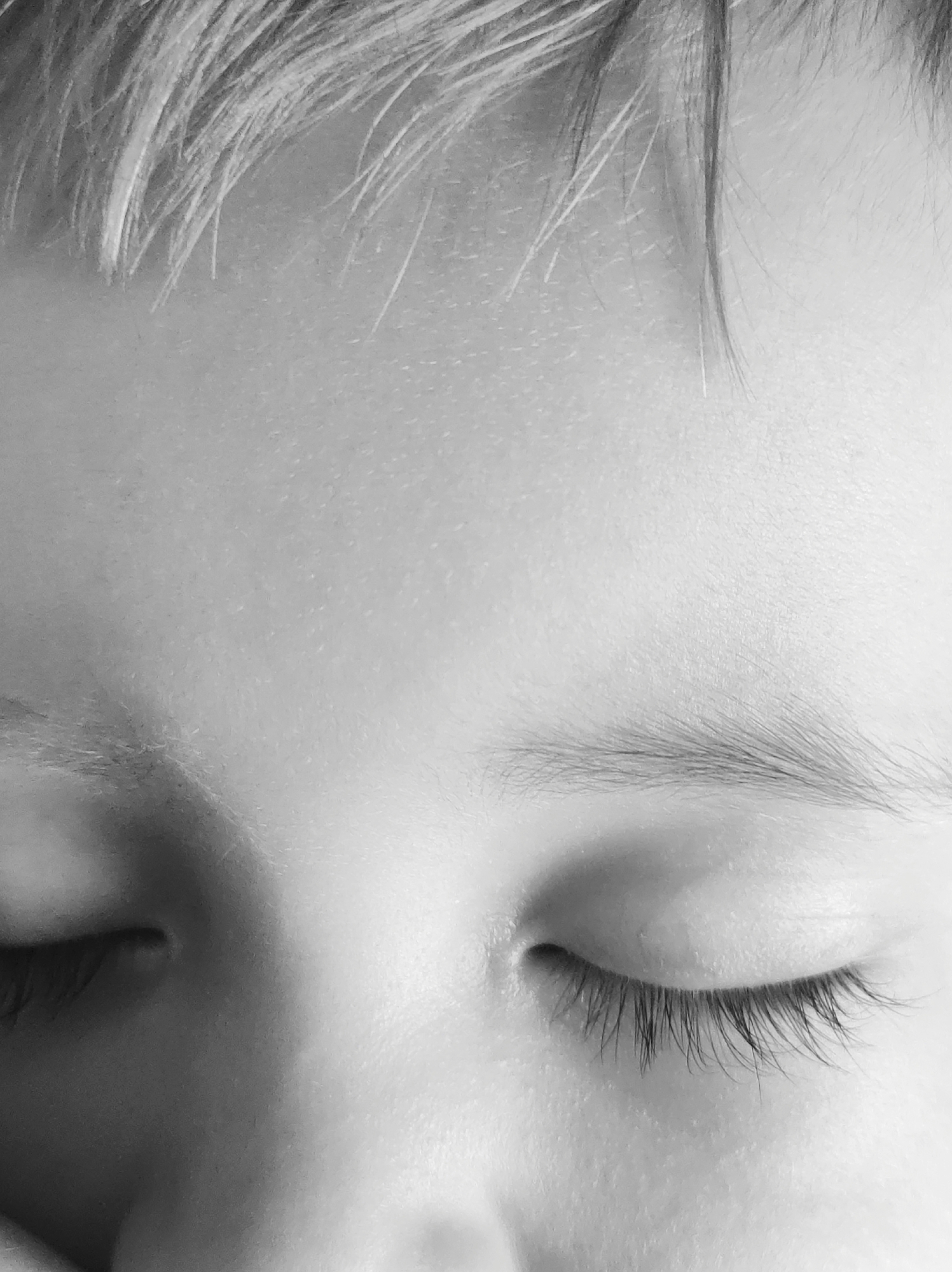 Close-up black and white image of a child's face with closed eyes. The child has light-colored hair. Eyebrows are visible, and eyelashes are prominent. The location is not discernible from the image.