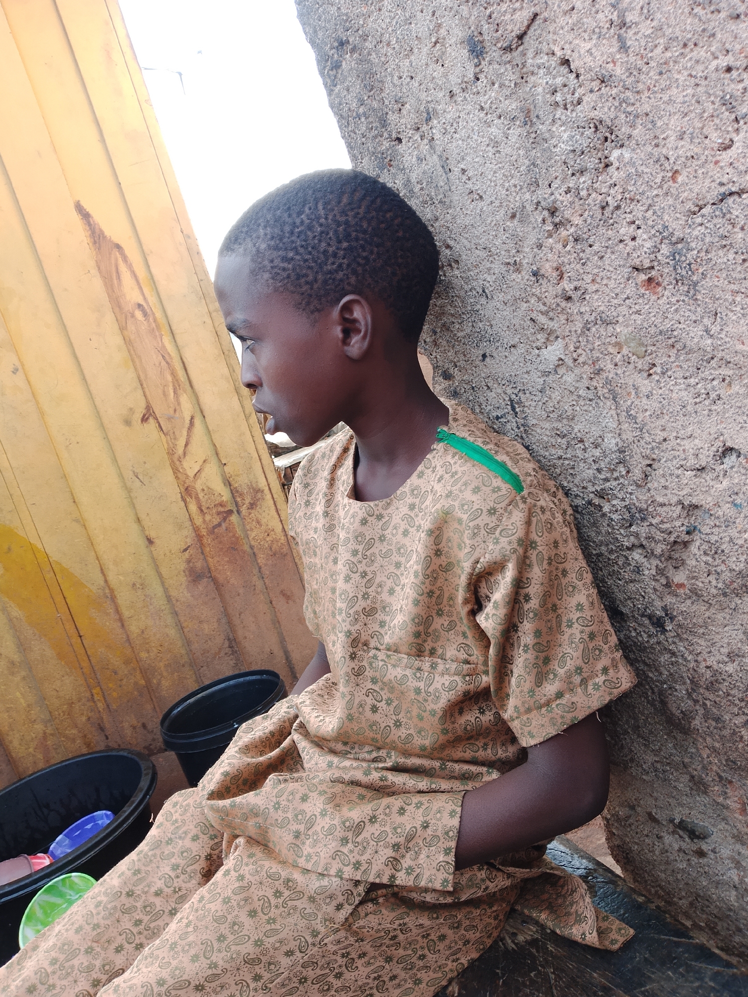 A young person with dark skin and short, textured hair sits leaning against a rough, textured concrete wall. They are wearing a light brown garment with a repeating paisley pattern in a darker shade of brown and green accents. A green stripe is visible on the left shoulder. To the left of the person, there is a yellow corrugated metal surface, likely part of a building or structure. In front of the person, on a dark surface, are two black plastic basins. The basin on the left contains a blue and a green plastic item, and possibly other items submerged in water. The basin on the right is empty. The environment appears to be outdoors, with a hint of a bright sky or light source in the upper left corner. The overall scene suggests a casual, possibly domestic, setting in Gura Top, Nigeria.