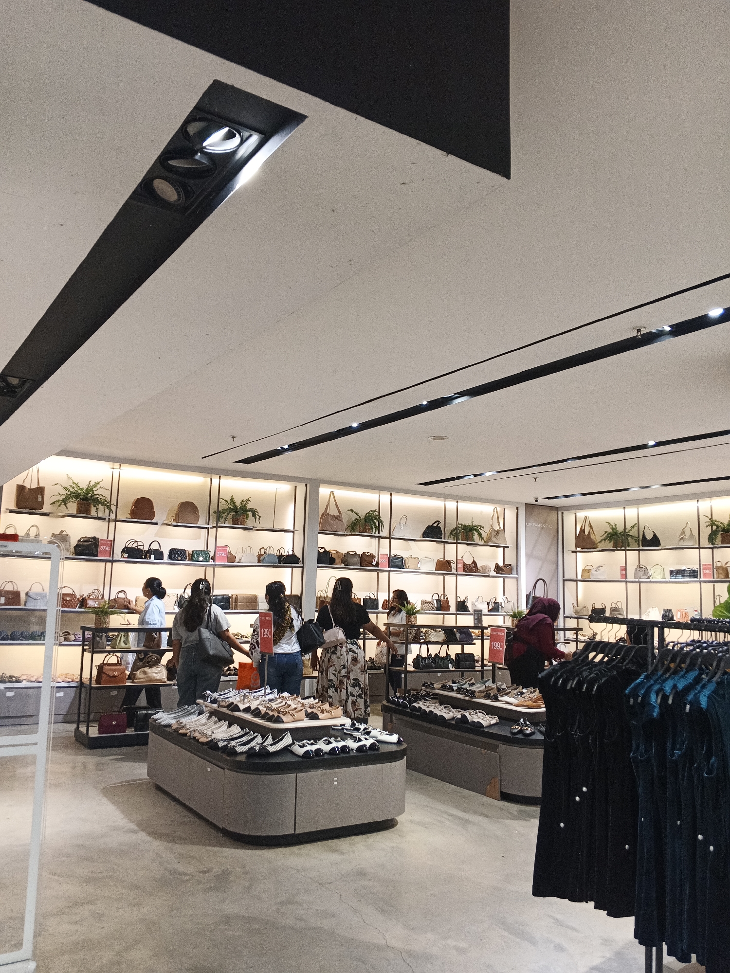 The image shows the interior of a well-lit retail store, likely a shoe and handbag boutique, in the city of Medan, Indonesia. The store features multiple shelves displaying an assortment of handbags and shoes. Several customers are browsing the merchandise. In the foreground, a display of shoes is arranged on a curved counter. Behind this, rows of shelves are filled with handbags of various sizes and colors, some with price tags visible. Plants are interspersed among the displays, adding a touch of greenery. The ceiling is a dominant feature, with black recessed lighting fixtures and dark structural elements. In the background, a rack of dark-colored clothing, possibly dresses or skirts, is visible. The store appears modern and organized, designed to showcase a wide range of fashion accessories.