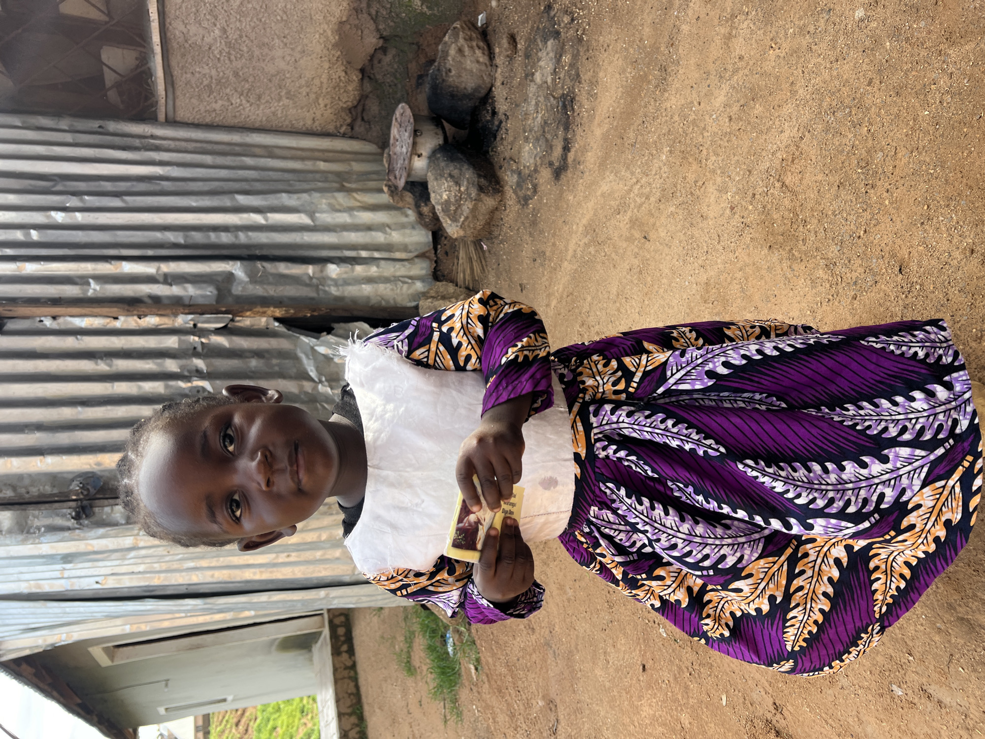 A young Black girl stands outdoors, holding a small object, possibly money, in her hands. She wears a white top and a vibrant purple and orange patterned skirt. Behind her, a corrugated metal structure leans against a rough, textured wall. The ground is covered in reddish-brown dirt, and in the background, a makeshift cooking area with rocks and a pot is visible. The environment suggests a rustic, possibly informal settlement.